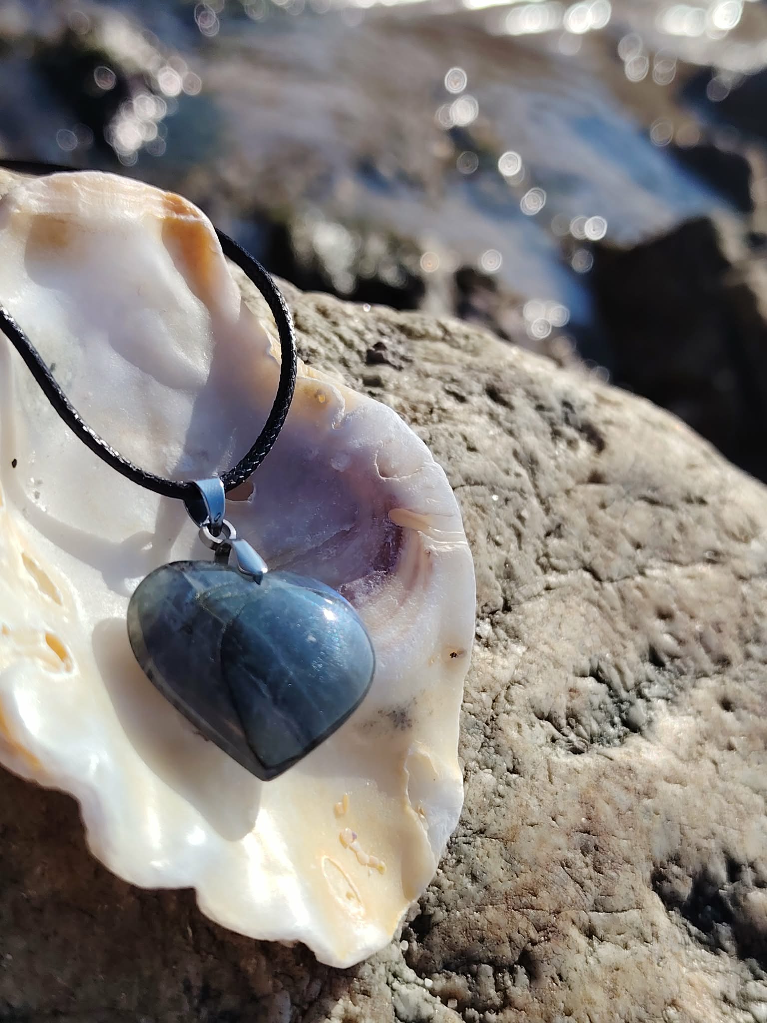 Pendentif cœur en labradorite sur cordon noir, présenté dans un coquillage sur rochers en Bretagne, collier pierre naturelle associé au réconfort, à l’intuition et à la paix intérieure.