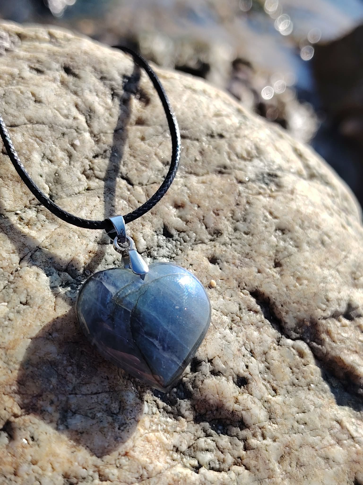 Pendentif cœur en labradorite sur cordon noir, posé sur les rochers en Bretagne, dans une ambiance minérale et lumineuse évoquant protection, intuition et apaisement
