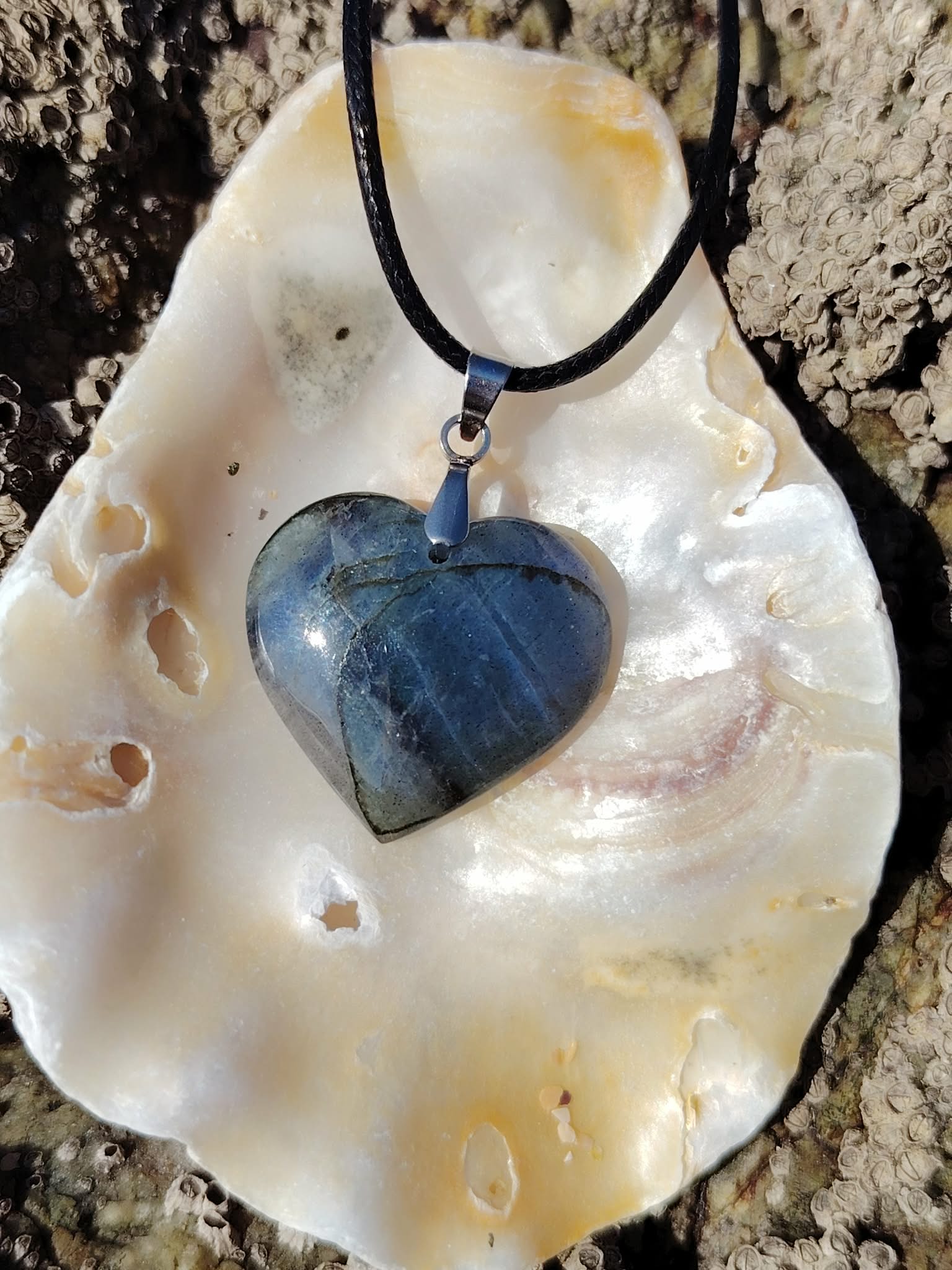 Pendentif cœur en labradorite posé sur un coquillage dans un décor minéral naturel, bijou aux reflets profonds associé à l’intuition, à la présence à soi et à la stabilité.