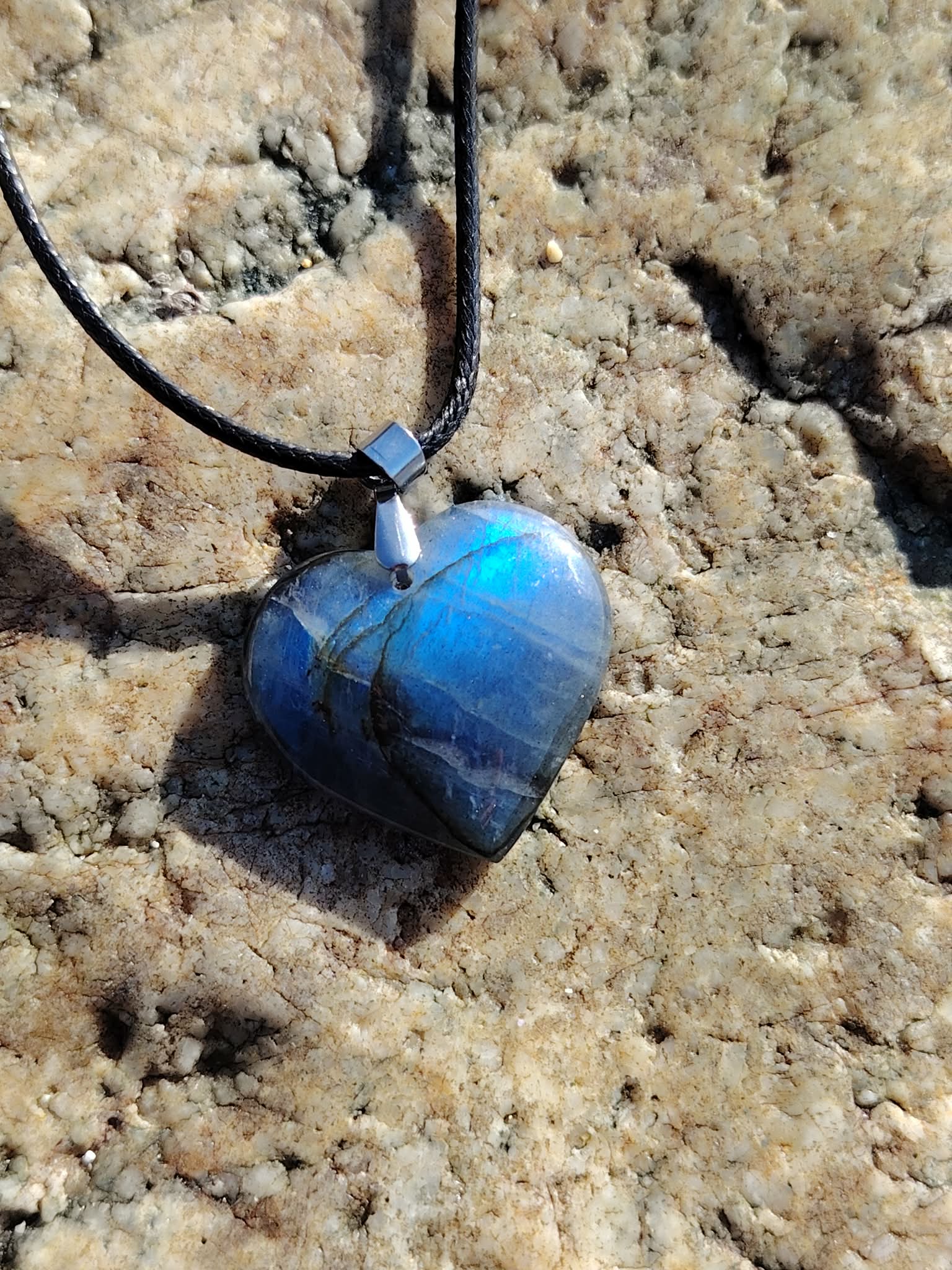 Pendentif cœur en labradorite sur cordon noir, posé sur rochers en Bretagne, bijou en pierre naturelle associé à l’intuition, à la stabilité intérieure et à la protection douce.