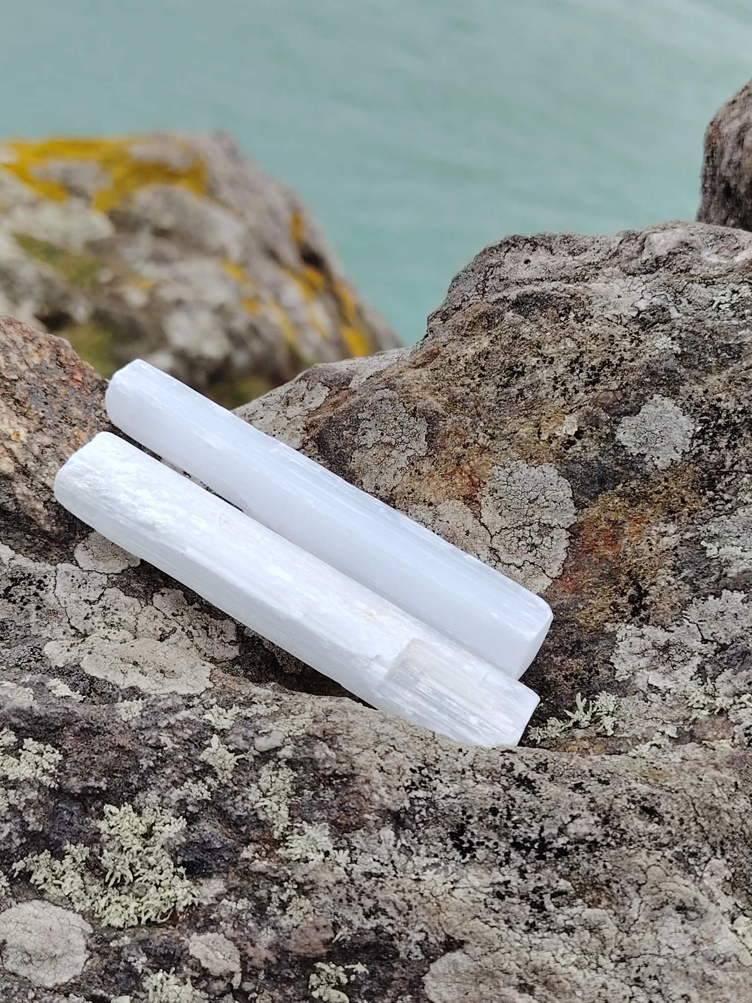 Deux baguettes de sélénite sur rochers en Bretagne sous ciel bleu, éclat nacré naturel, douceur intérieure et énergie limpide.
