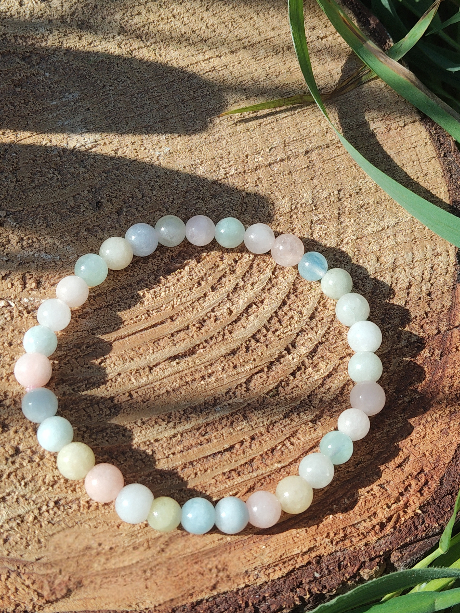 Bracelet Morganite 6 mm sur tranche de bois au soleil avec brins d’herbe, perles pastel aux nuances douces, bijou en pierre naturelle évoquant la tranquillité, l’amour de soi et l’apaisement émotionnel.