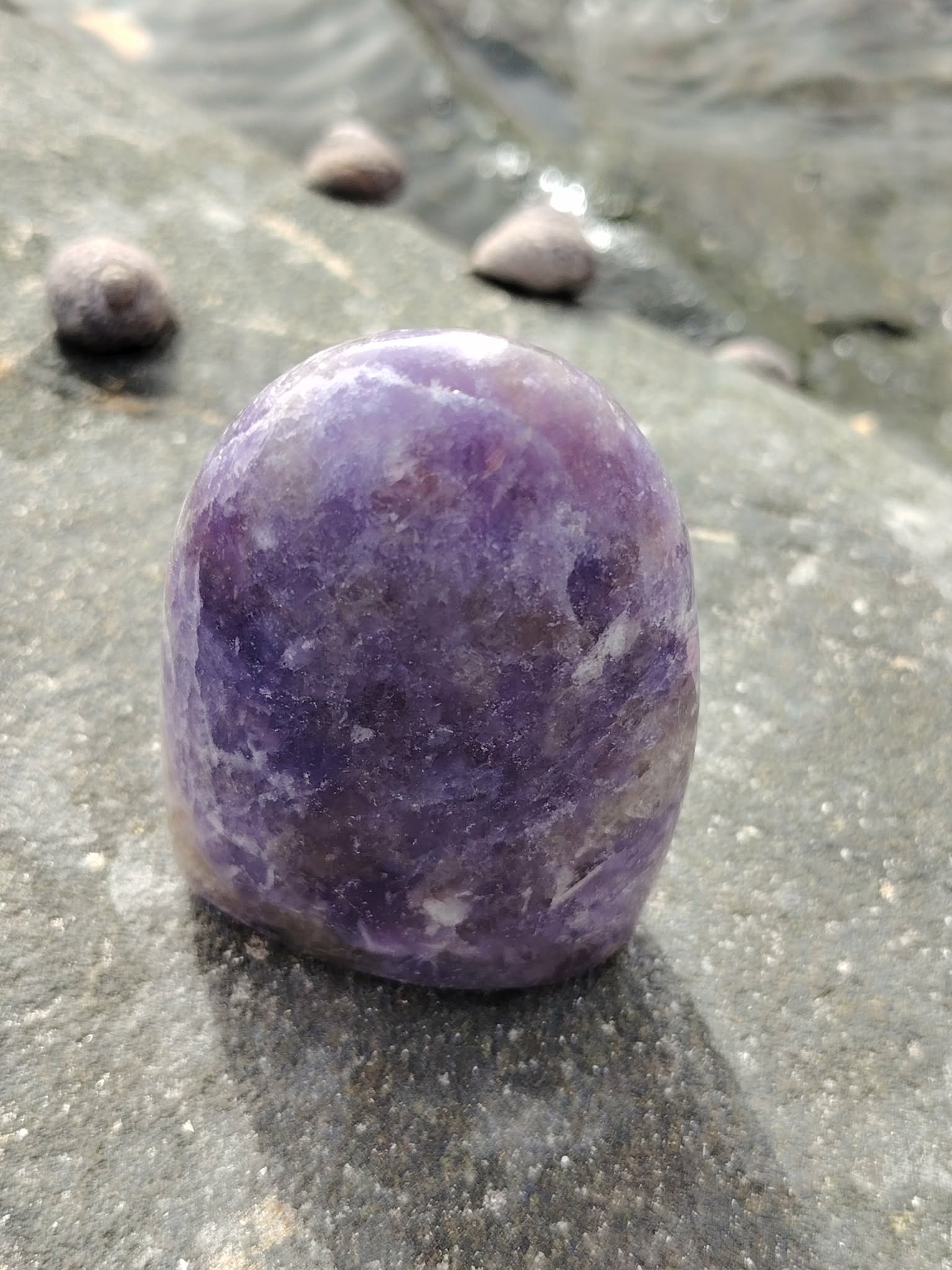 Lépidolite forme libre violette posée sur pierre humide près de l’eau, pierre naturelle évoquant l’apaisement, la souplesse émotionnelle et la sérénité.