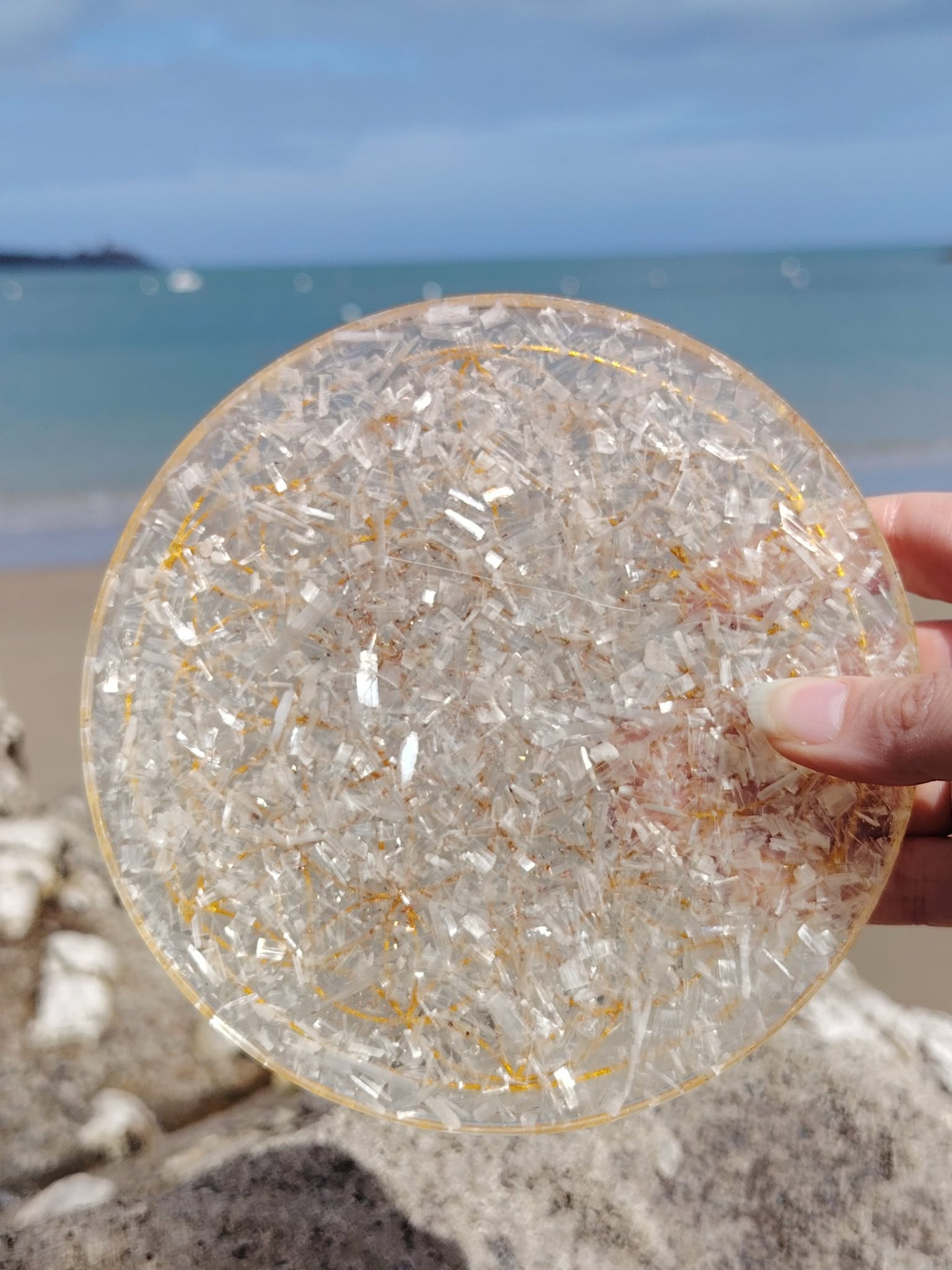 Plateau de rechargement orgonite sélénite photographié sur plage en Bretagne avec roche au premier plan, disque blanc lumineux et énergie apaisante, harmonie émotionnelle.