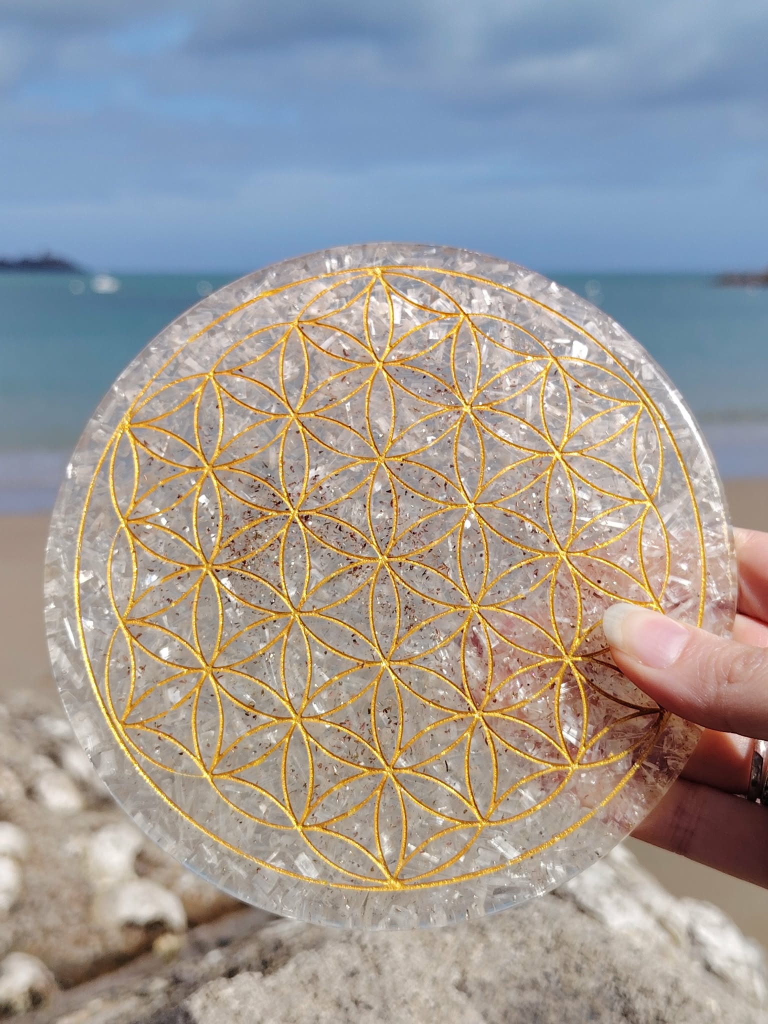 Plateau orgonite sélénite tenu en main face à la mer en Bretagne, disque blanc aux reflets nacrés et symbole Fleur de Vie, sérénité intérieure et équilibre vibratoire.