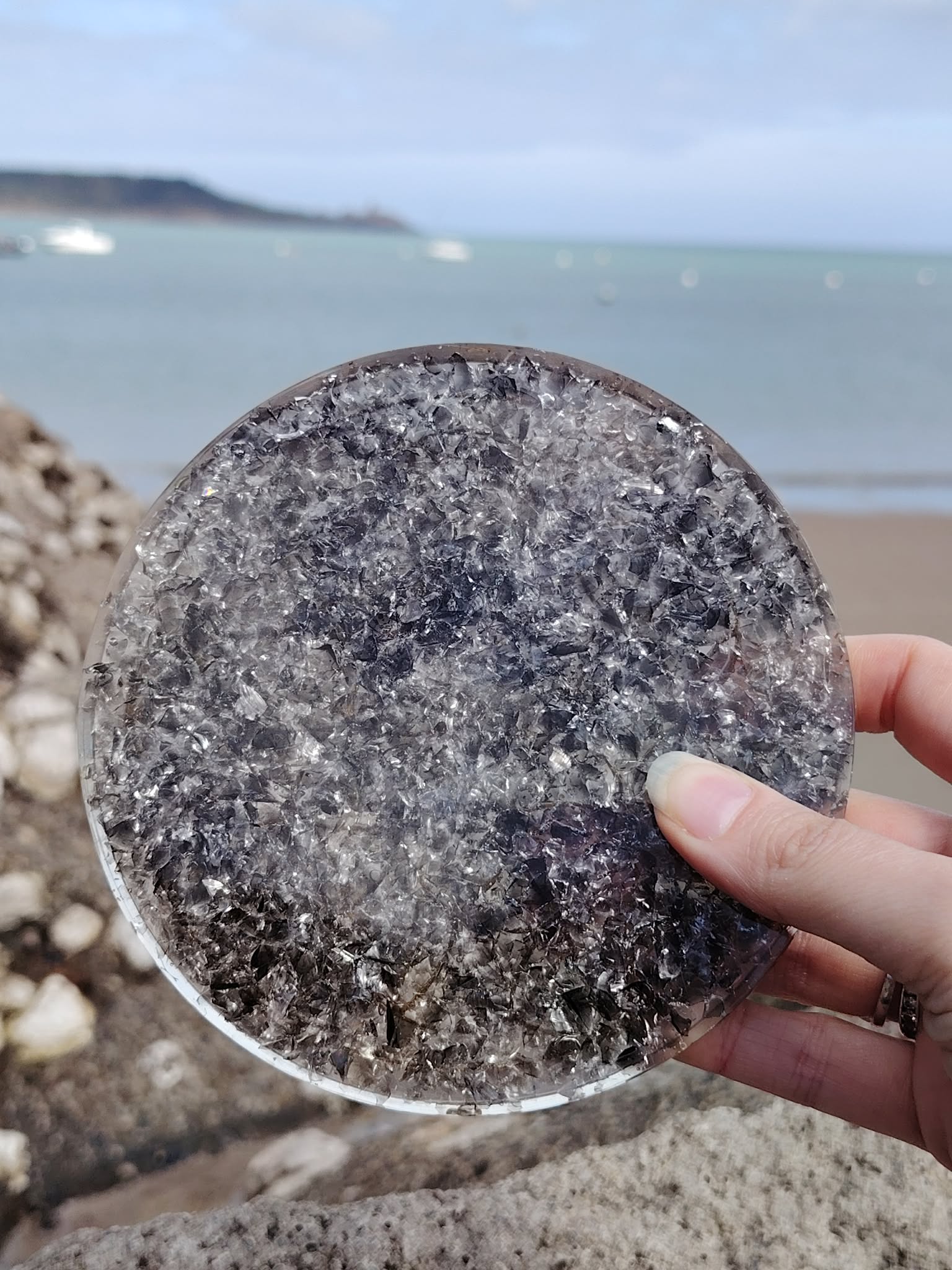 Plateau orgonite obsidienne noire tenu en main face à la mer en Bretagne, disque minéral gris noir poli, protection vibratoire et recentrage émotionnel.