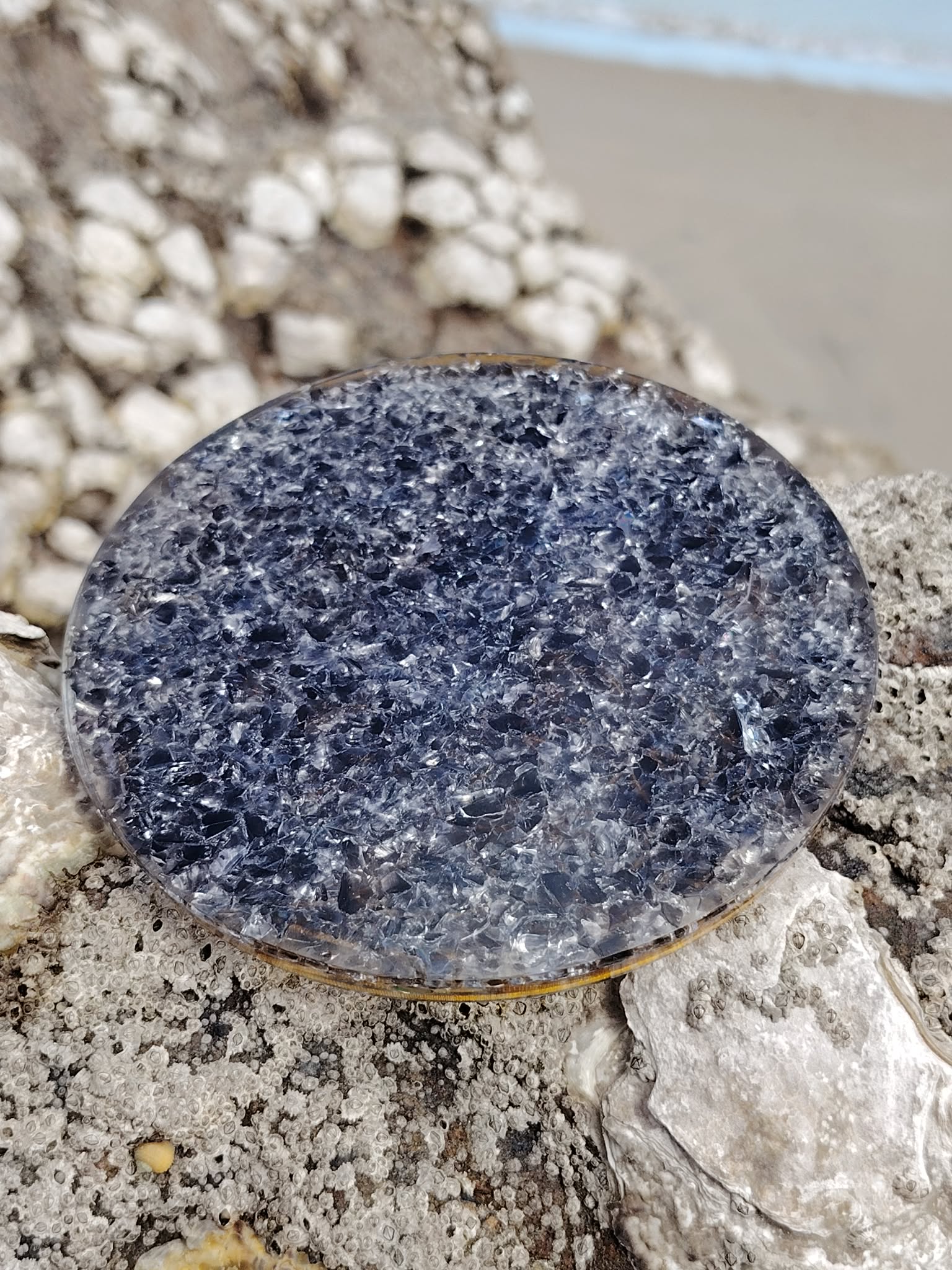 Plateau orgonite obsidienne noire sur rocher marin en Bretagne avec plage floue en fond, face minérale sombre sans motif visible, équilibre intérieur et énergie stabilisante.