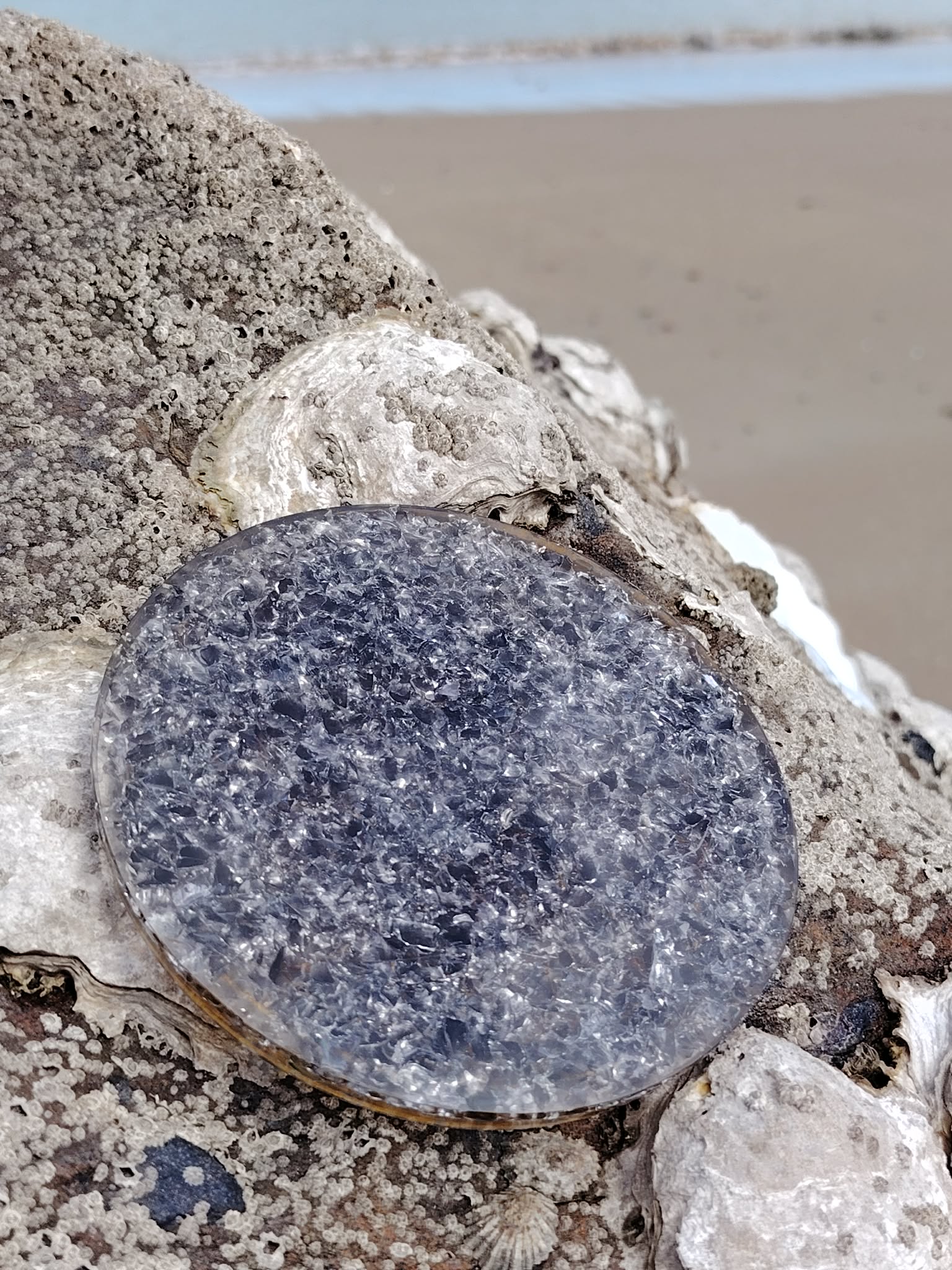 Plateau de rechargement orgonite obsidienne noire posé sur roche marine en Bretagne avec plage en arrière-plan, face minérale gris noir, énergie protectrice et retour au calme.