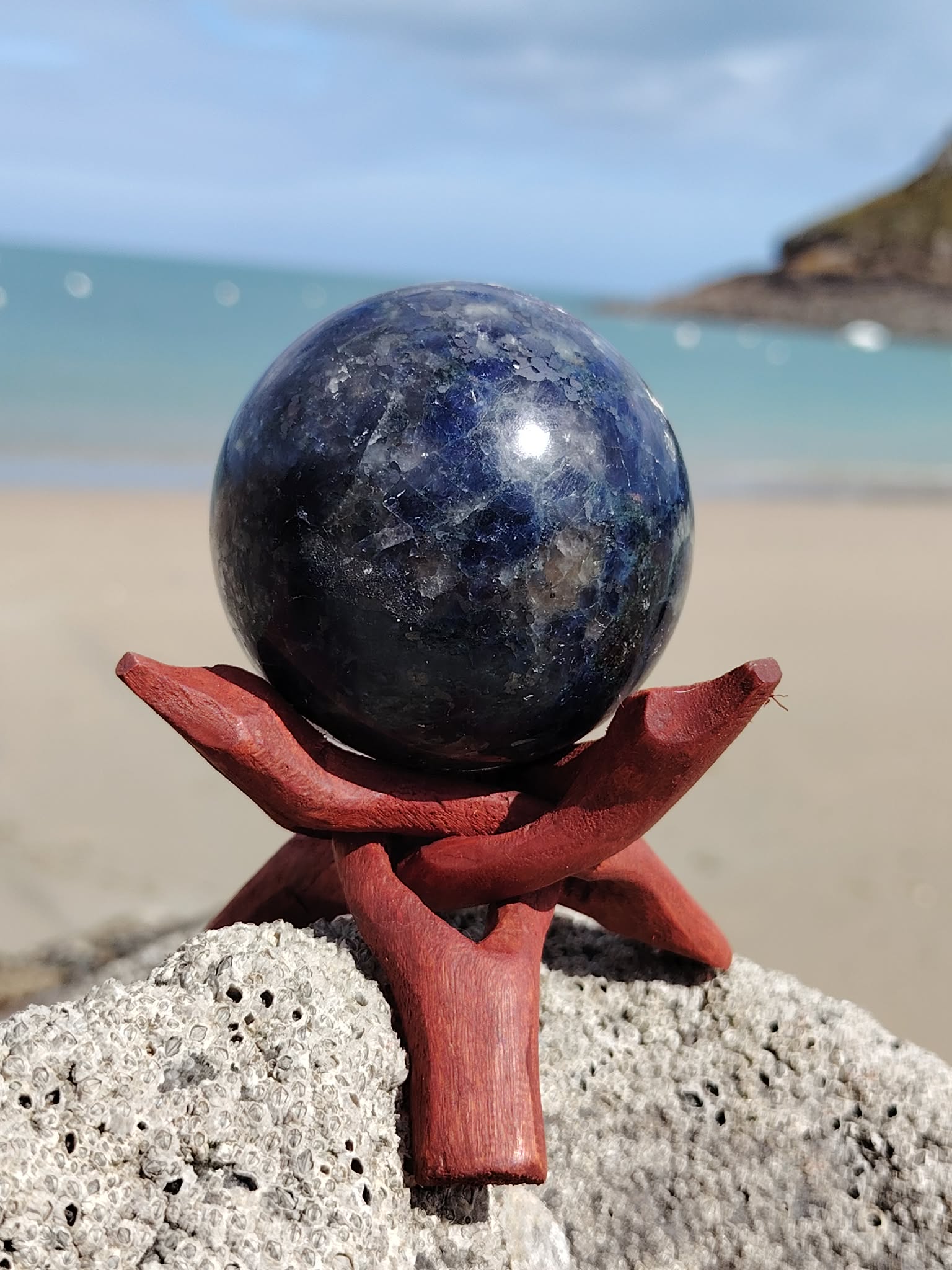 Sphère iolite ou cordiérite sur roche marine en Bretagne avec socle en bois, pierre bleu nuit aux éclats nuancés, sérénité mentale et intuition apaisée.