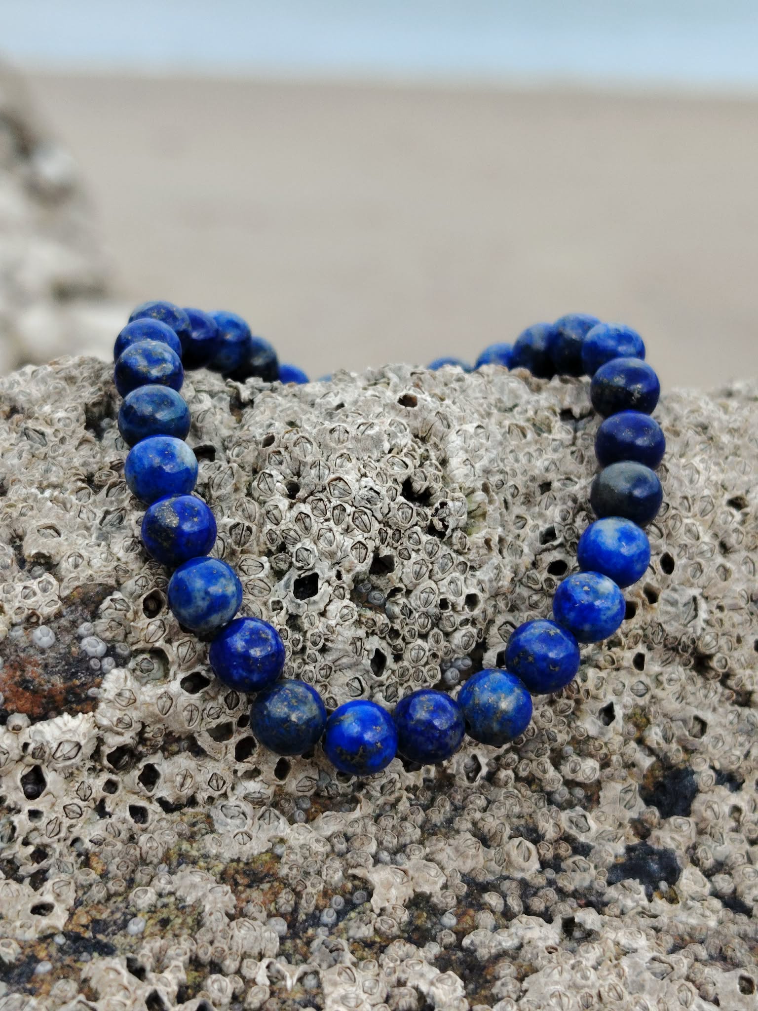 Bracelet Lapis Lazuli 6 mm formant un cœur sur roche de bord de mer, perles bleu intense aux touches dorées, vérité intérieure et harmonie émotionnelle.