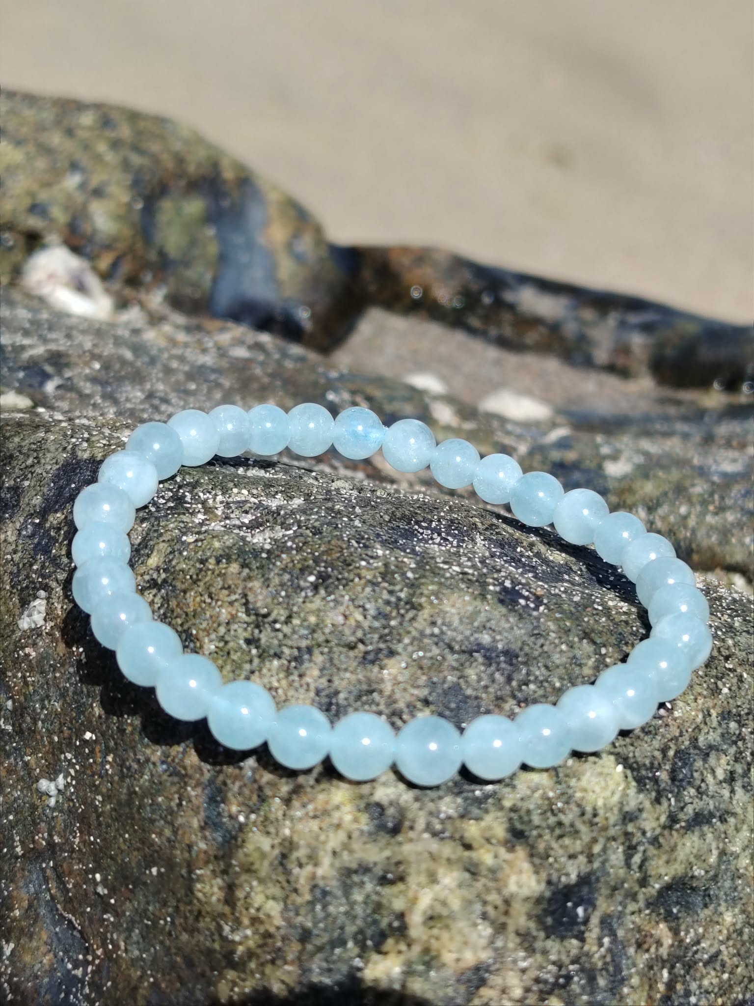 Bracelet Aigue-Marine 6 mm posé sur rochers en bord de mer, perles bleu lagon lumineuses, sérénité intérieure et communication douce.