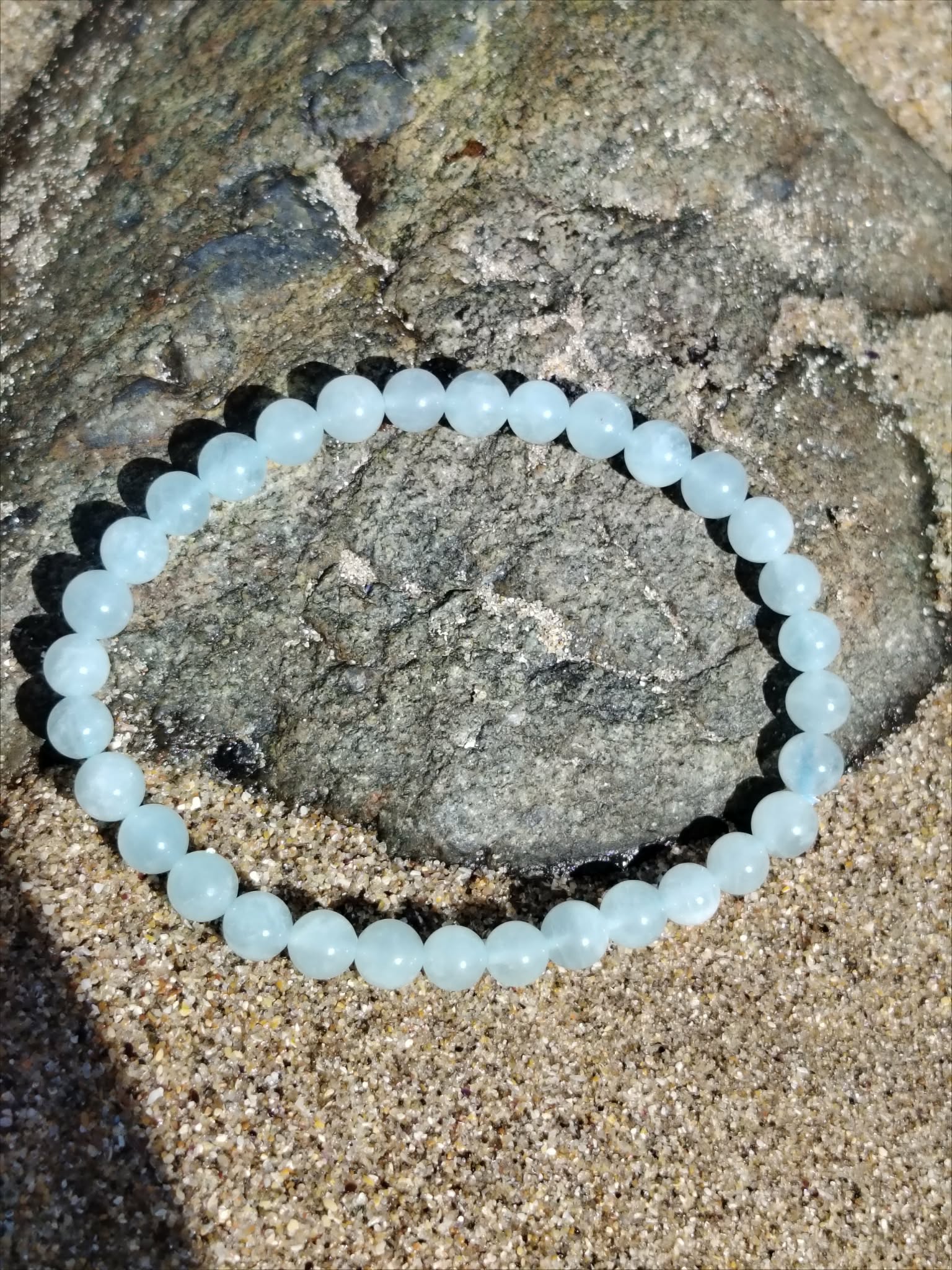 Bracelet Aigue-Marine 6 mm sur sable près d’un rocher côtier, perles bleu tendre, calme mental et recentrage émotionnel.