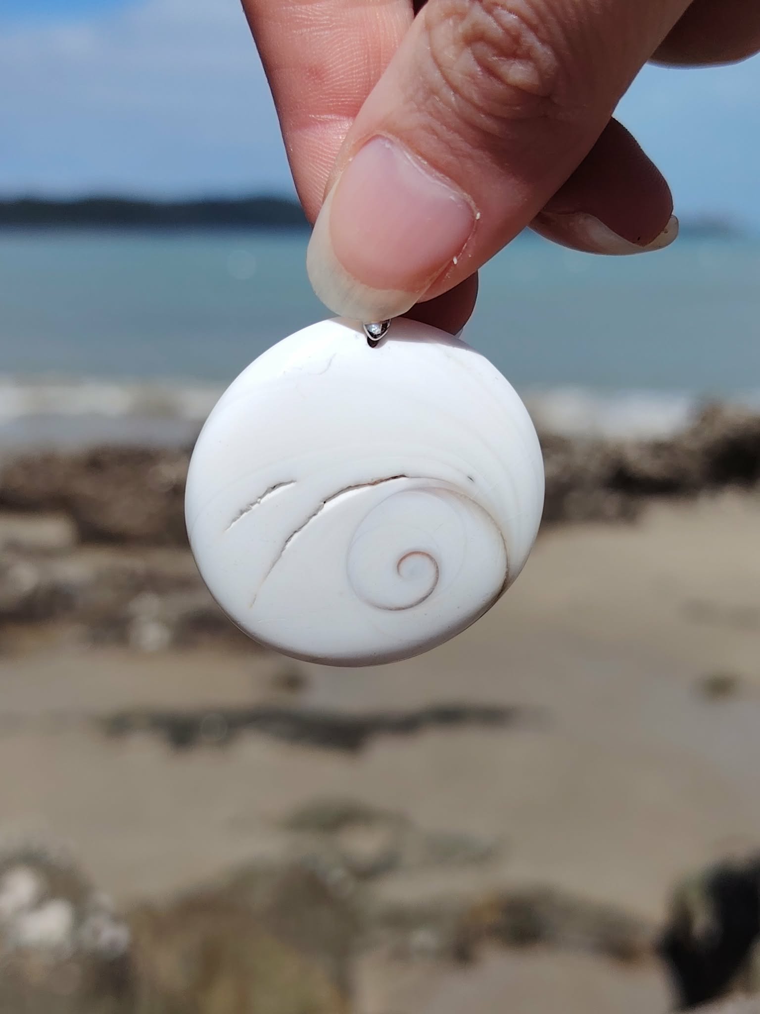 Pendentif œil de Sainte Lucie blanc présenté à la main devant la plage et l’horizon marin, coquillage naturel à spirale beige, paix intérieure et chance.