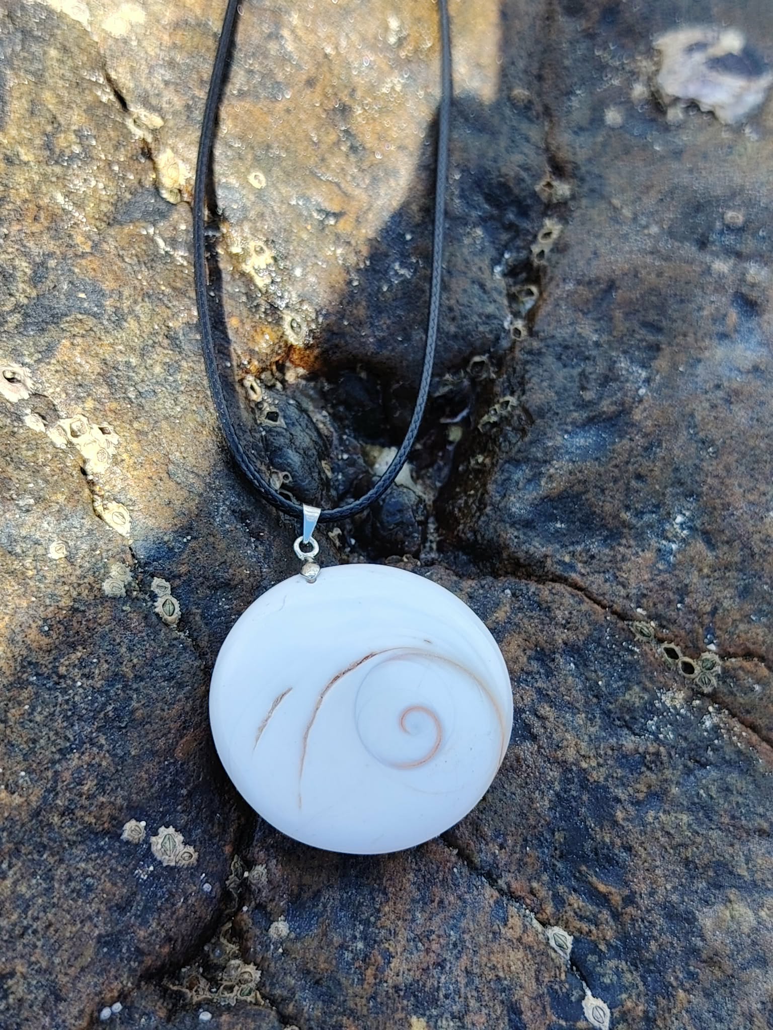 Pendentif rond œil de Sainte Lucie blanc sur lien noir, photographié sur roche sombre en extérieur, coquillage naturel associé à l’intuition et à la protection.