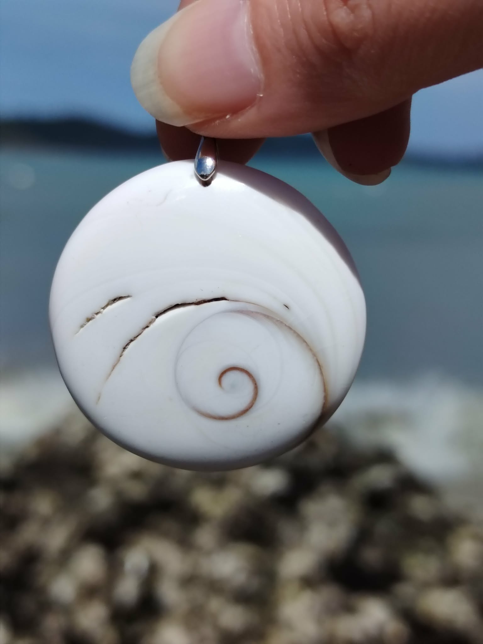 Pendentif œil de Sainte Lucie rond blanc tenu entre les doigts, spirale beige visible sur fond marin, coquillage porte-bonheur lié à la chance et au calme intérieur.