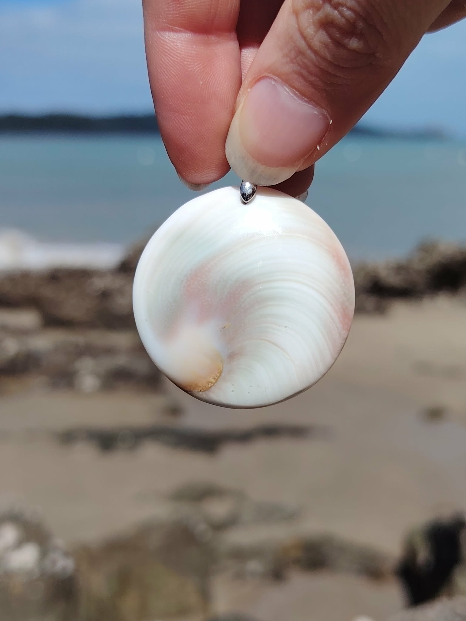 Face nacrée d’un pendentif œil de Sainte Lucie rond blanc tenue entre les doigts, sur fond de plage et horizon marin, sérénité et énergie apaisante.