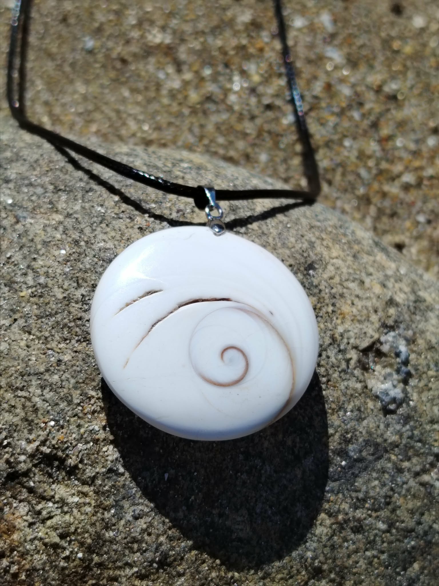 Pendentif œil de Sainte Lucie rond blanc sur cordon noir, posé sur roche granitique en lumière naturelle, coquillage marin porte-bonheur aux reflets doux, symbole de protection et d’intuition.