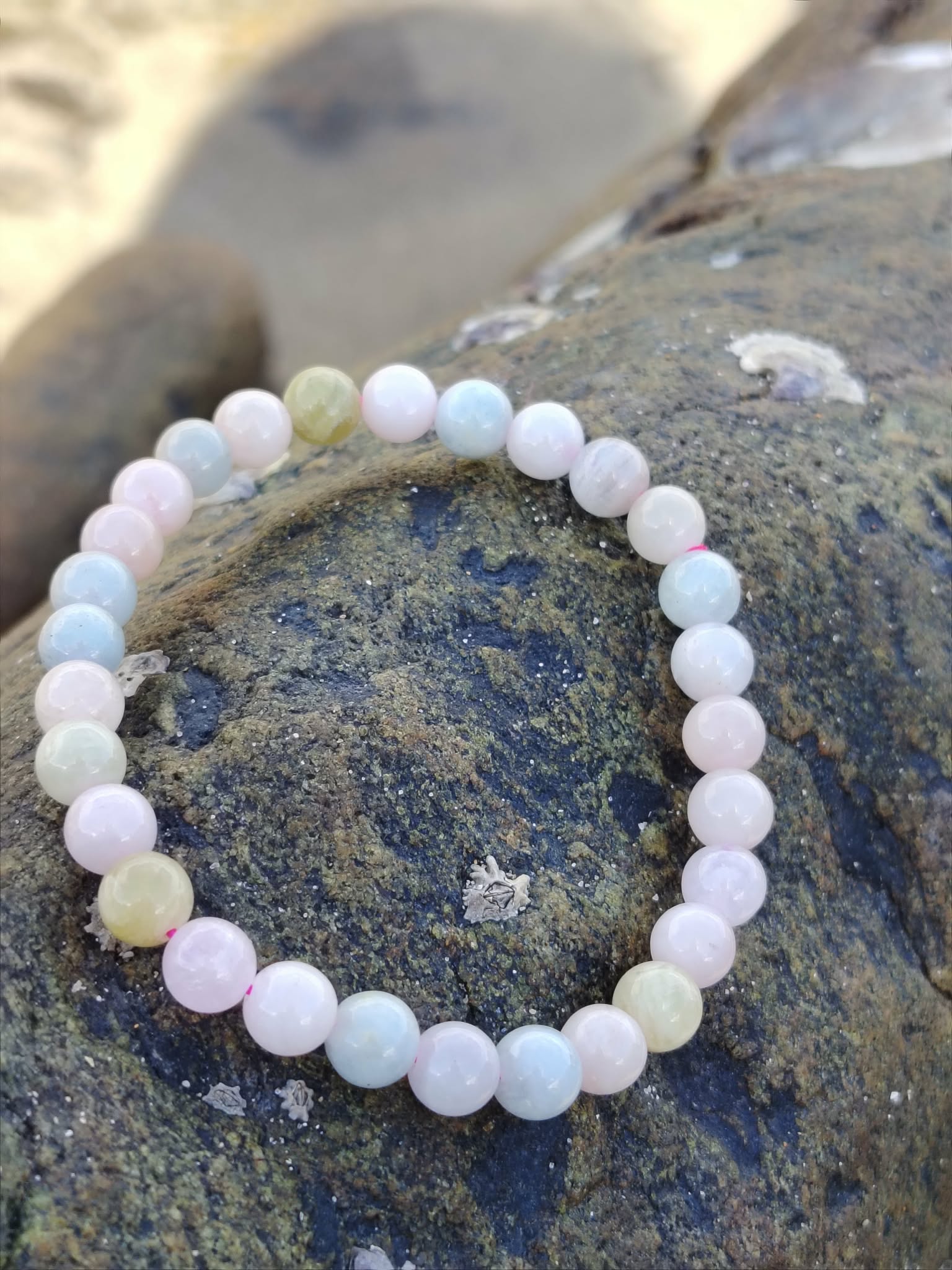 Bracelet Morganite 6 mm photographié sur roche bretonne au bord de l’eau, perles roses, bleues et vert pâle, bijou en pierre naturelle évoquant la sérénité, la délicatesse et l’équilibre affectif.