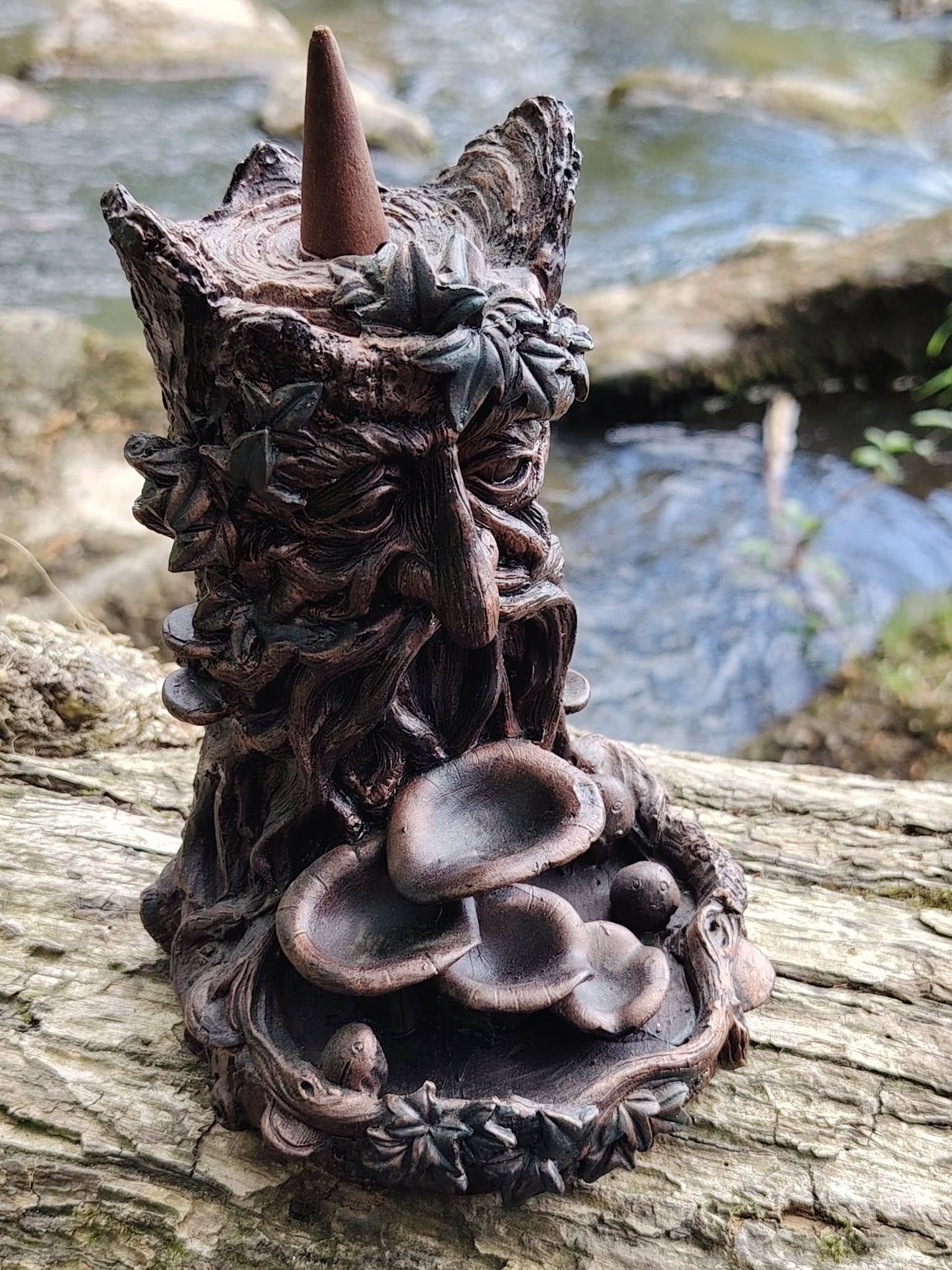 Brûleur d’encens à refoulement arbre en résine bronze posé sur bois en forêt avec ruisseau en arrière-plan, visage sculpté, coupelles en cascade et cône d’encens sur le dessus.