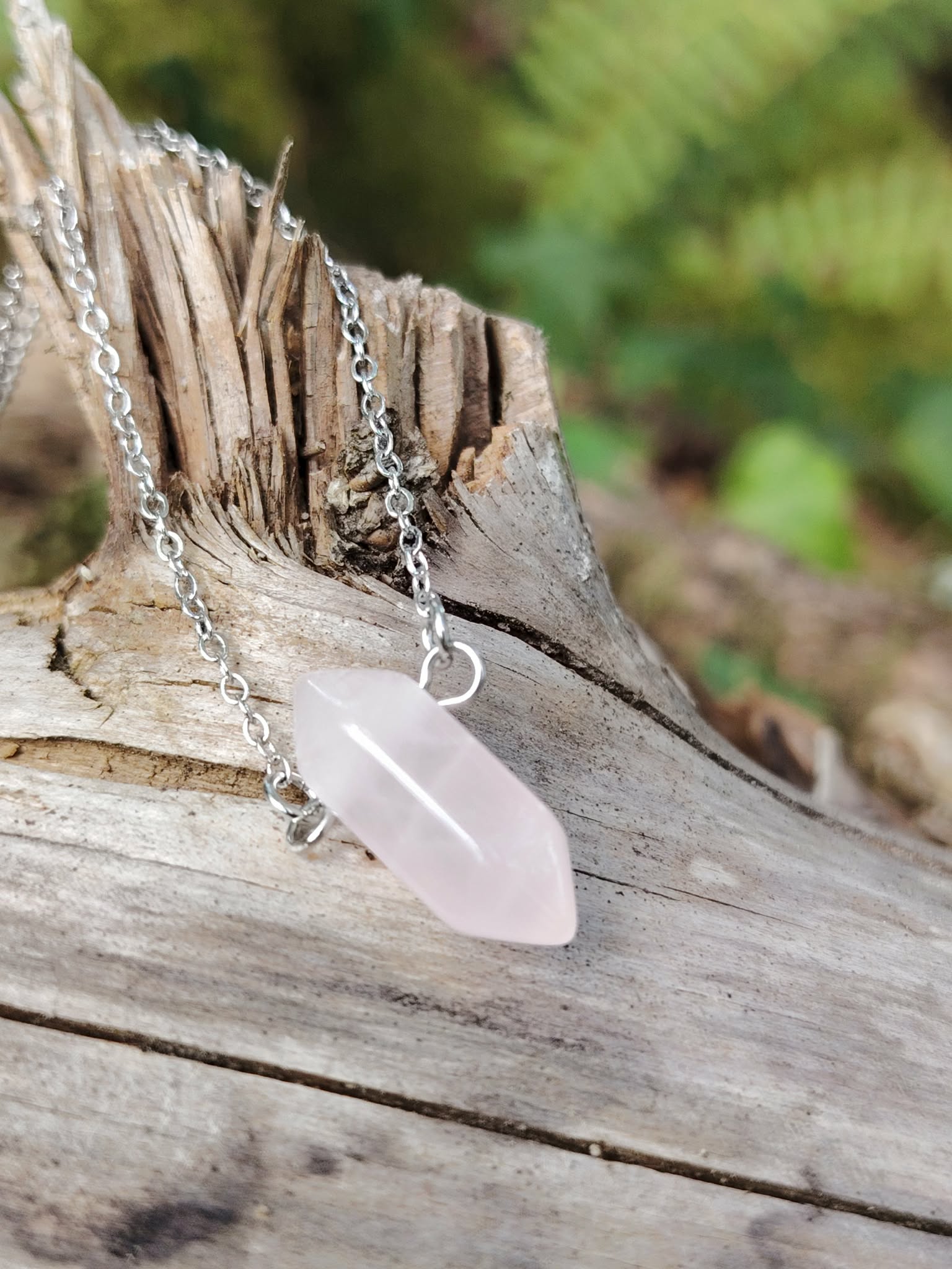 Pendentif pointe en quartz rose sur chaîne argentée posé sur bois brut fissuré, pierre rose délicate avec fond végétal flou, évoquant douceur relationnelle, rééquilibrage affectif et paix intérieure.