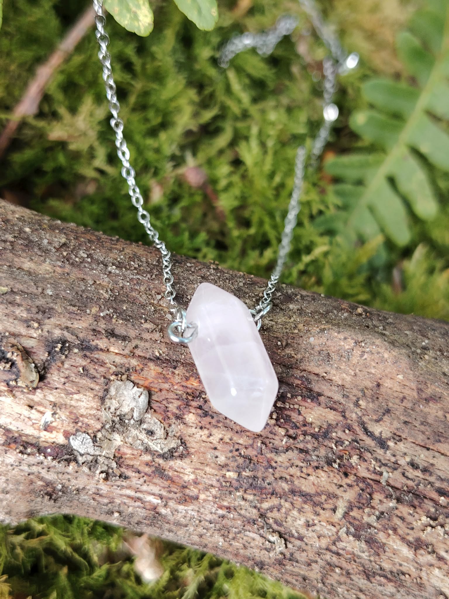 Pendentif pointe en quartz rose sur chaîne argentée, pierre rose pâle posée sur une branche avec mousse verte, symbole de douceur, apaisement émotionnel et ouverture du cœur.