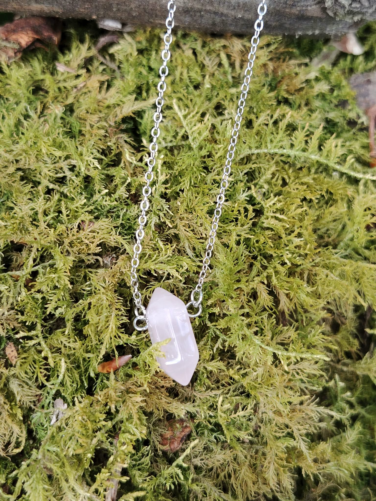 Pendentif pointe en quartz rose sur chaîne argentée suspendu sous une branche, pierre rose posée sur mousse végétale, évoquant apaisement du cœur, sérénité émotionnelle et douceur affective.