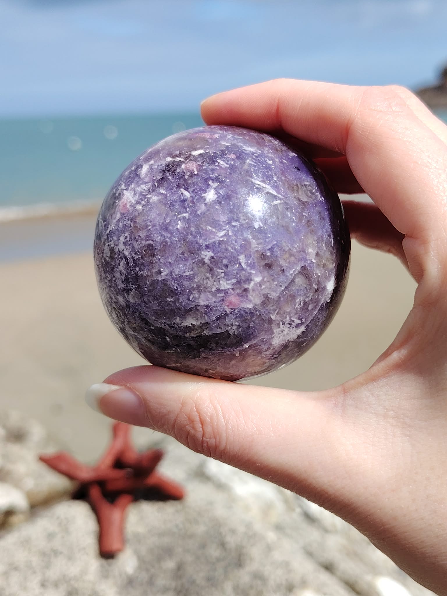 Sphère lépidolite tenue entre les doigts au bord de mer en Bretagne, pierre violette aux marbrures naturelles, apaisement du mental et recentrage.