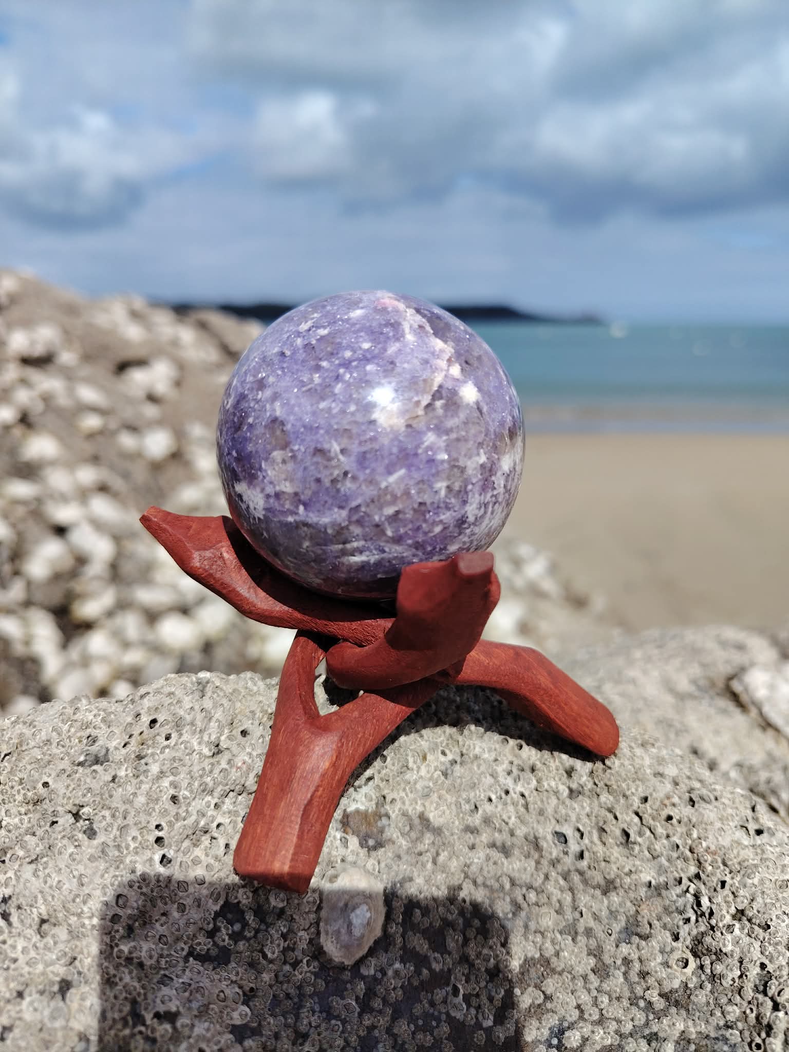 Sphère lépidolite photographiée sur rocher au bord de mer en Bretagne avec support en bois, pierre violette naturelle, sérénité intérieure et équilibre émotionnel.