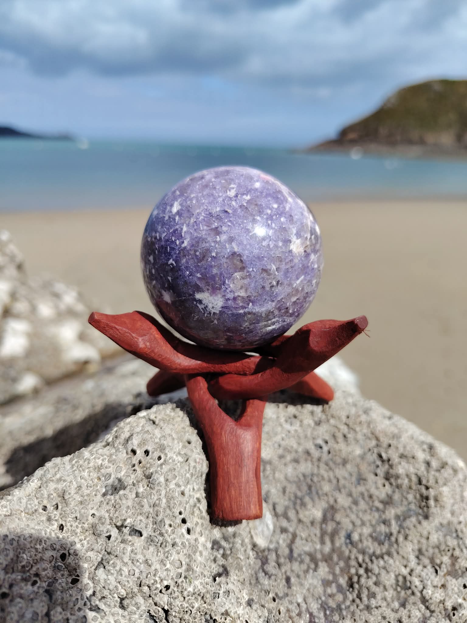 Sphère lépidolite sur roche marine en Bretagne face à la plage avec support en bois, pierre lavande naturelle, détente émotionnelle et énergie apaisante.