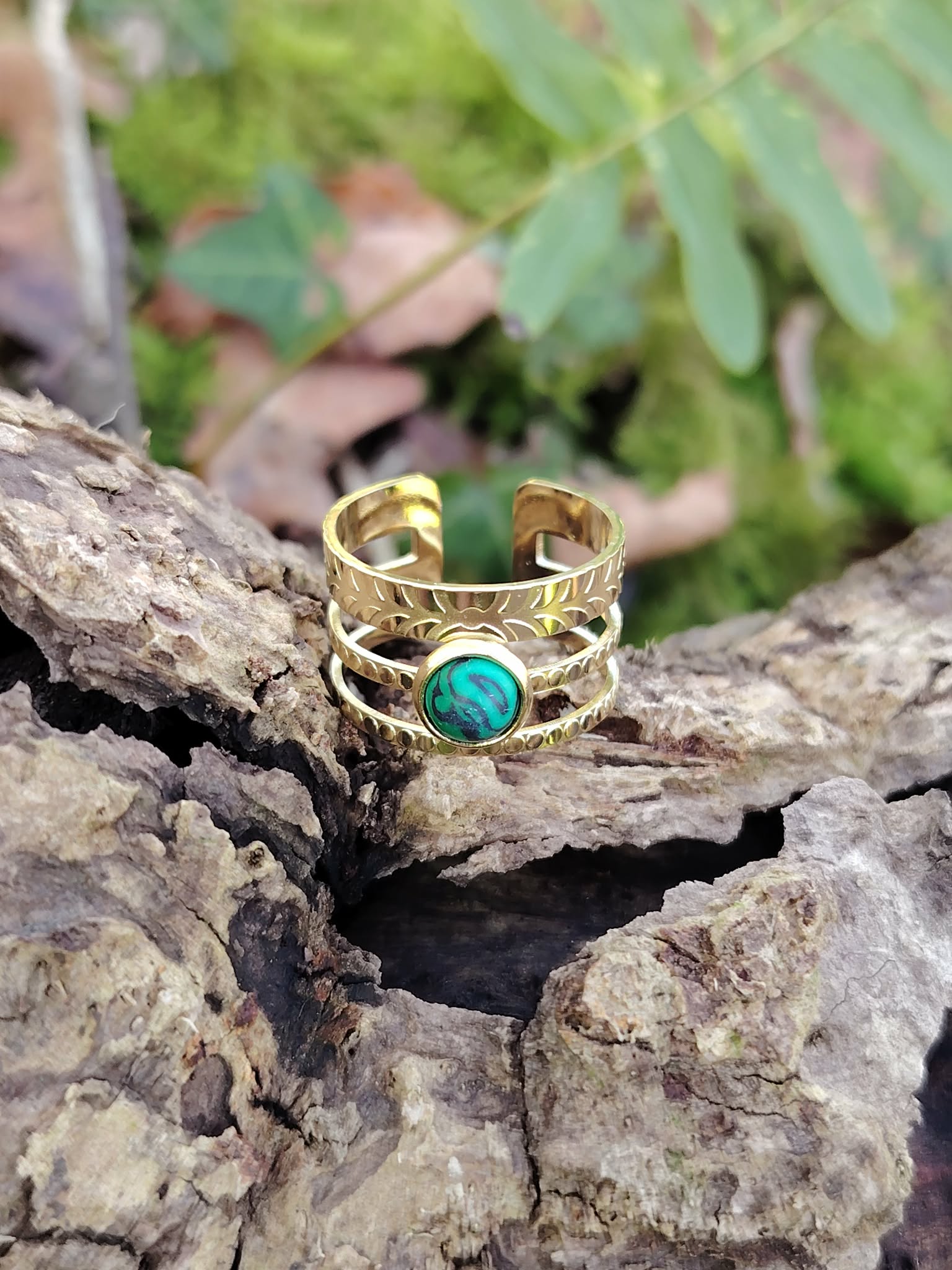 Bague dorée ajustable en acier inoxydable posée dans un tronc creux en forêt, pierre verte ovale au visuel malachite, douceur intérieure et équilibre émotionnel.