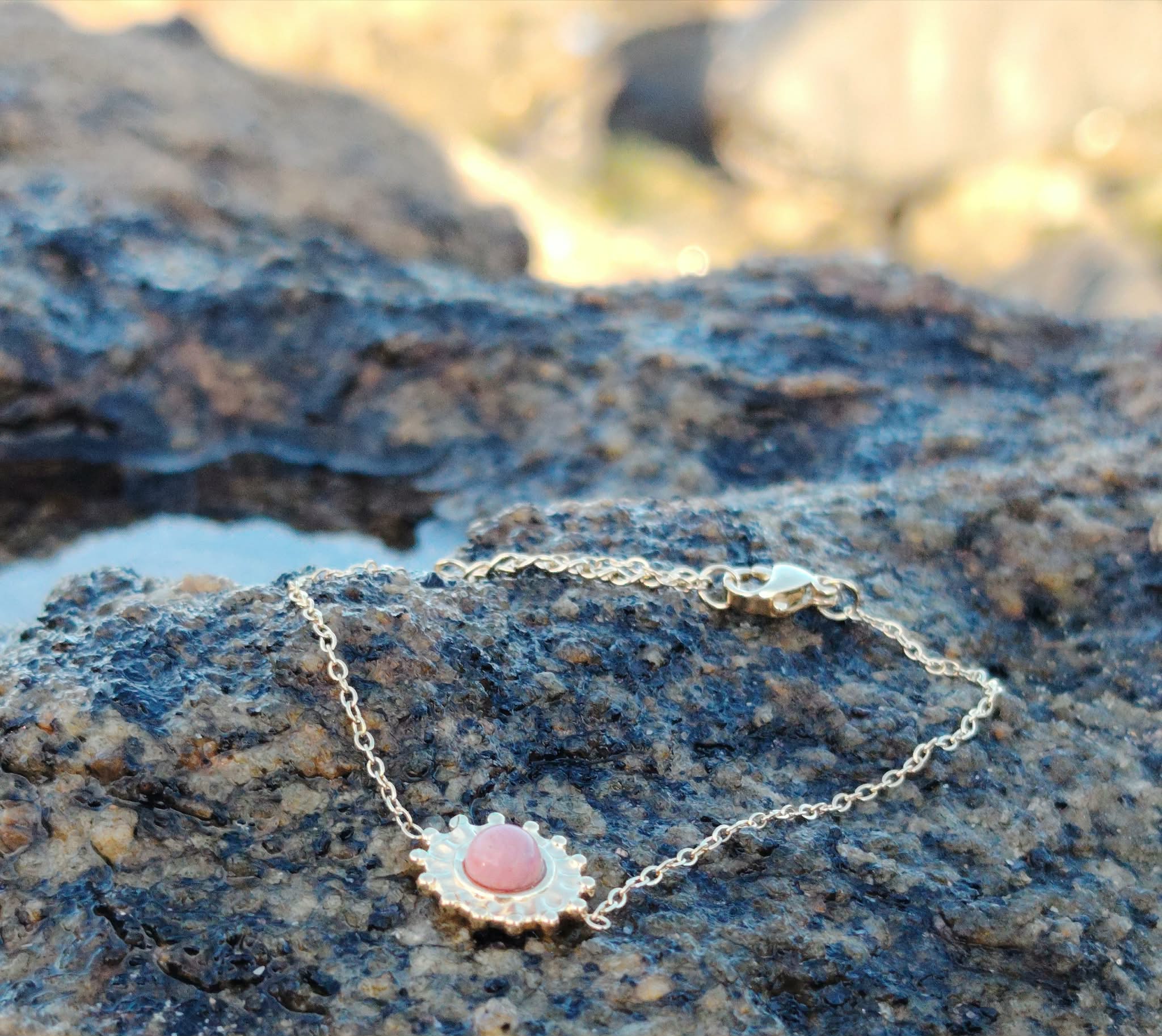 Bracelet soleil doré en acier inoxydable avec rhodonite rose posé sur roche en bord de mer en Bretagne, motif solaire délicat, harmonie intérieure et style raffiné.