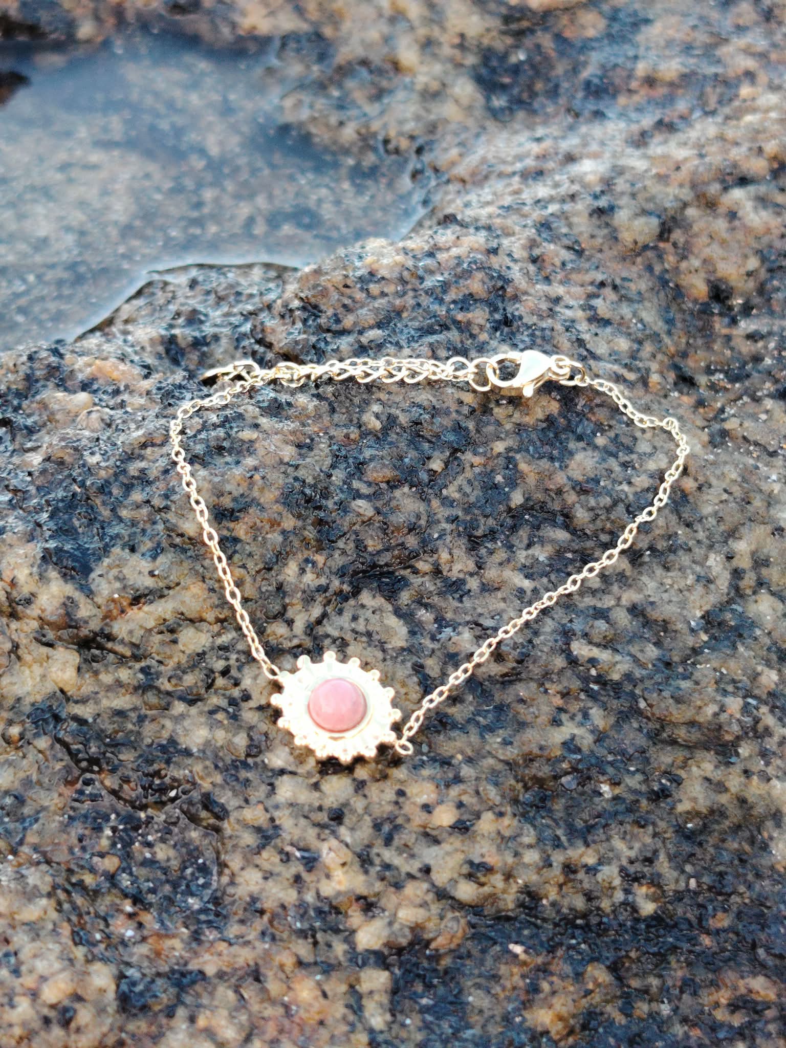 Bracelet fin doré avec soleil et rhodonite naturelle sur rocher coquillé face à la mer en Bretagne, acier inoxydable brillant et pierre rose tendre, charme délicat et énergie apaisante.