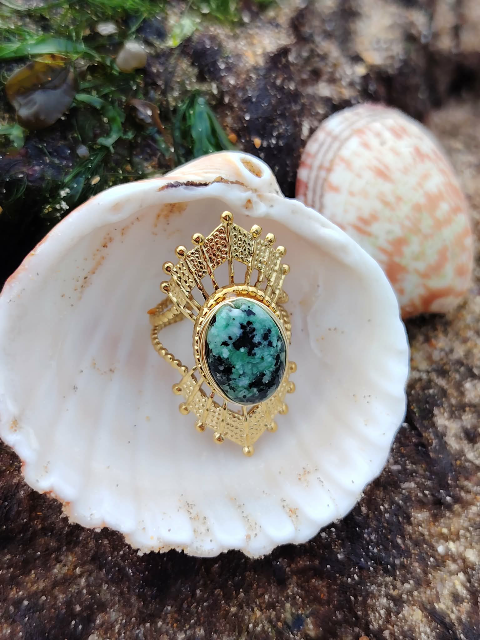 Bague dorée ajustable solaire avec pierre verte ovale sur coquillage au bord de mer en Bretagne, acier inoxydable lumineux, élégance naturelle et harmonie intérieure.