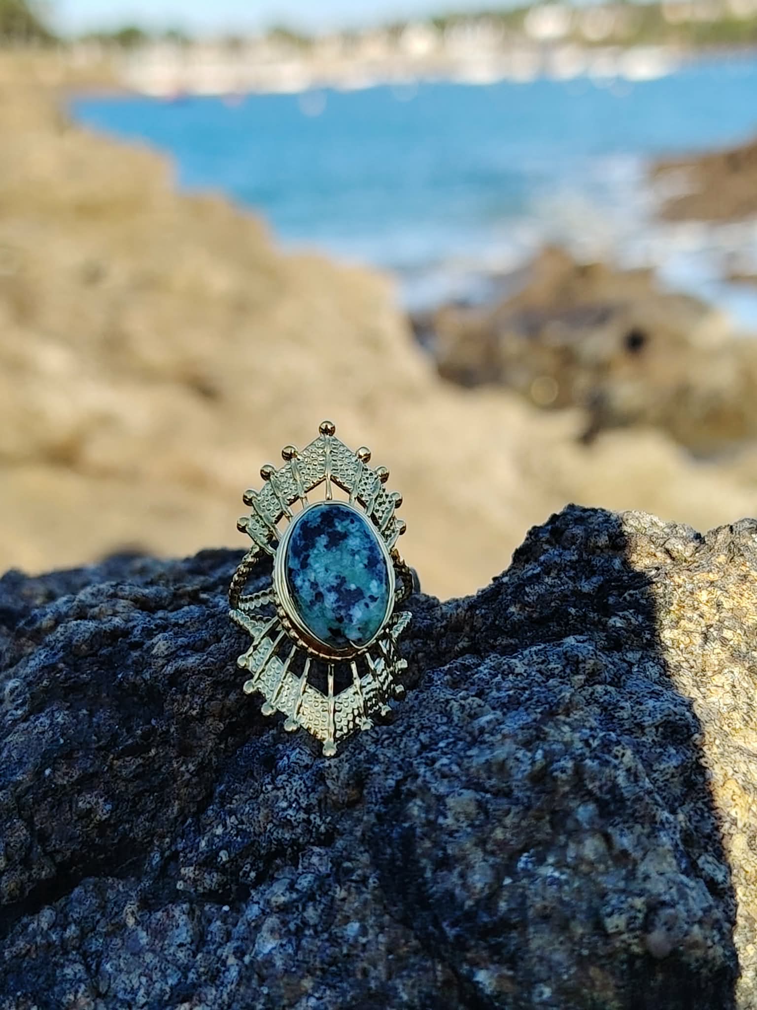 Bague dorée ajustable solaire avec pierre verte ovale sur rocher face à la mer en Bretagne, acier inoxydable brillant, énergie apaisante et allure élégante.