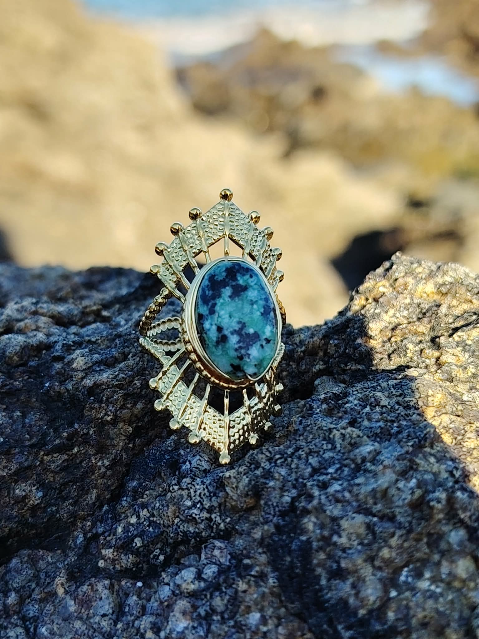 Bague solaire dorée ajustable avec pierre verte ovale sur roche côtière en Bretagne, acier inoxydable brillant et cabochon vert profond, style raffiné et équilibre émotionnel.