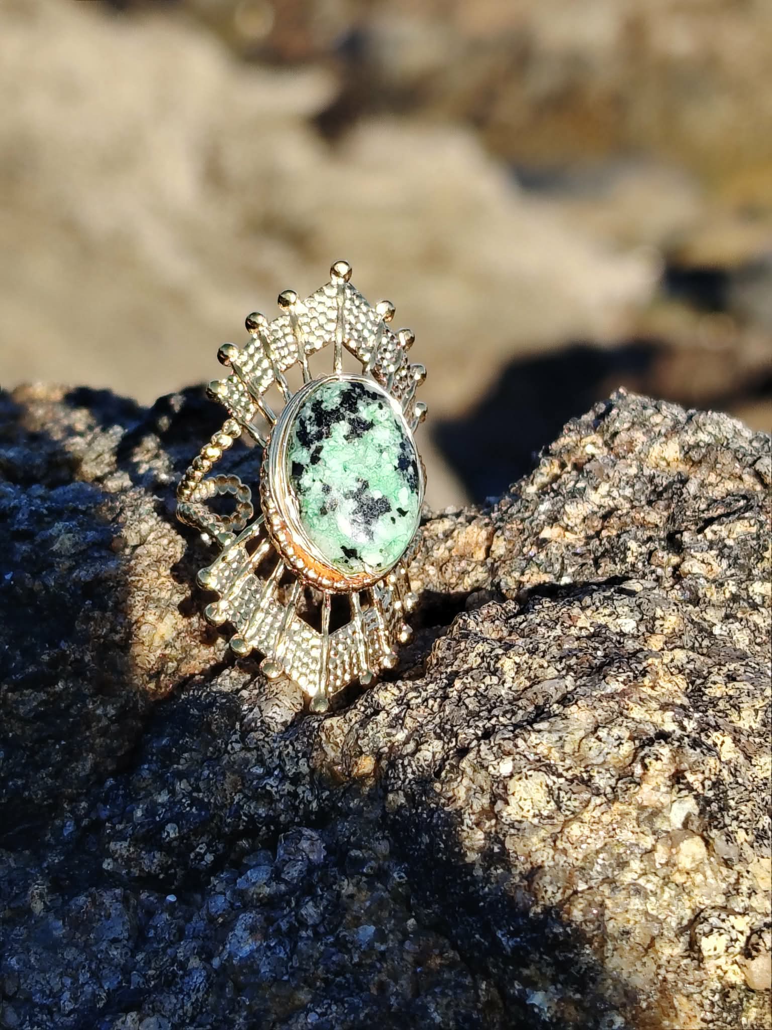 Bague solaire dorée en acier inoxydable sur roche marine en Bretagne, pierre verte ovale aux nuances profondes, élégance lumineuse et stabilité émotionnelle.