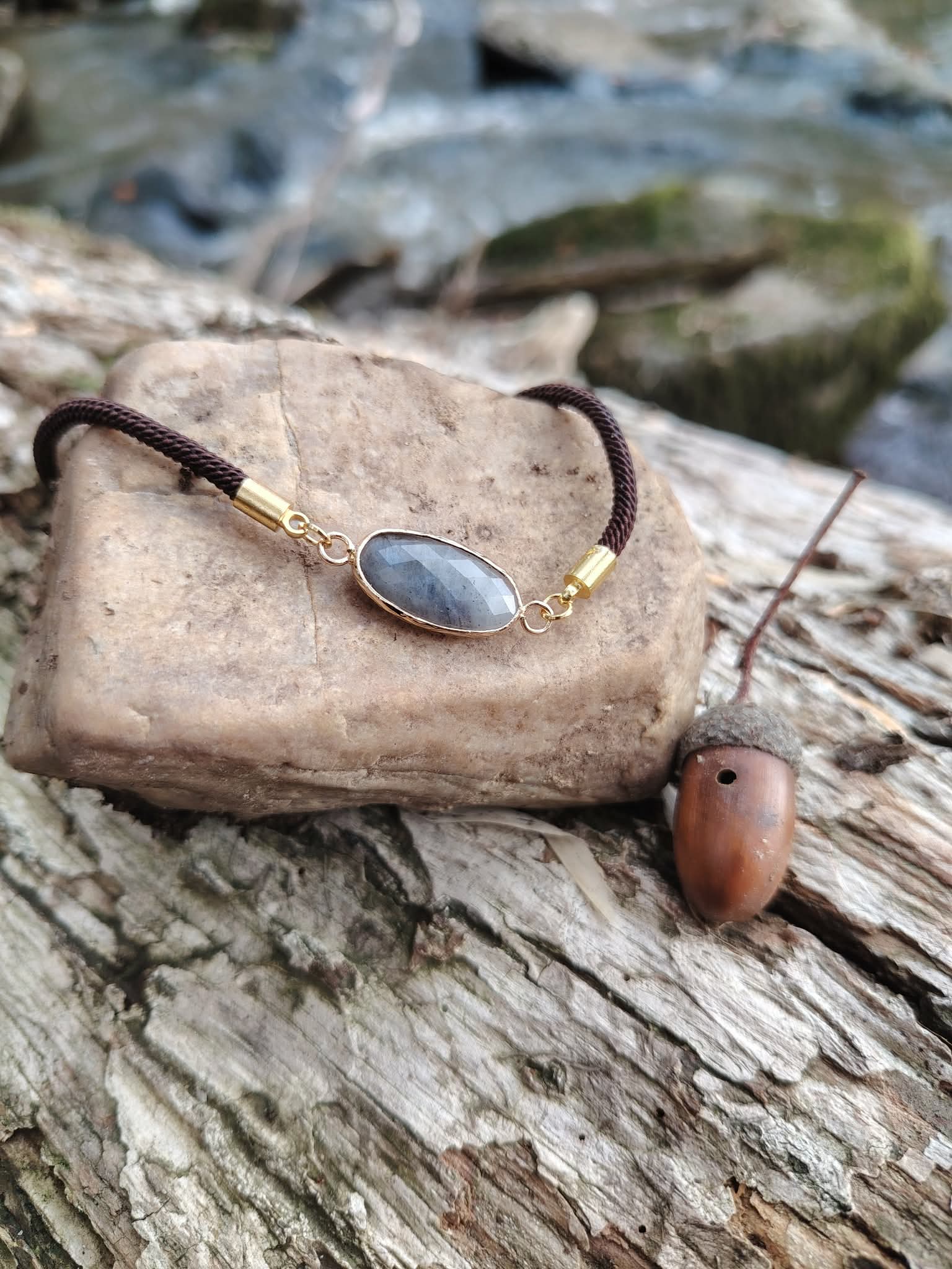 Bracelet labradorite facettée présenté sur bois naturel avec gland près d’un cours d’eau, médaillon ovale doré et cordon brun, recentrage et douceur intérieure.