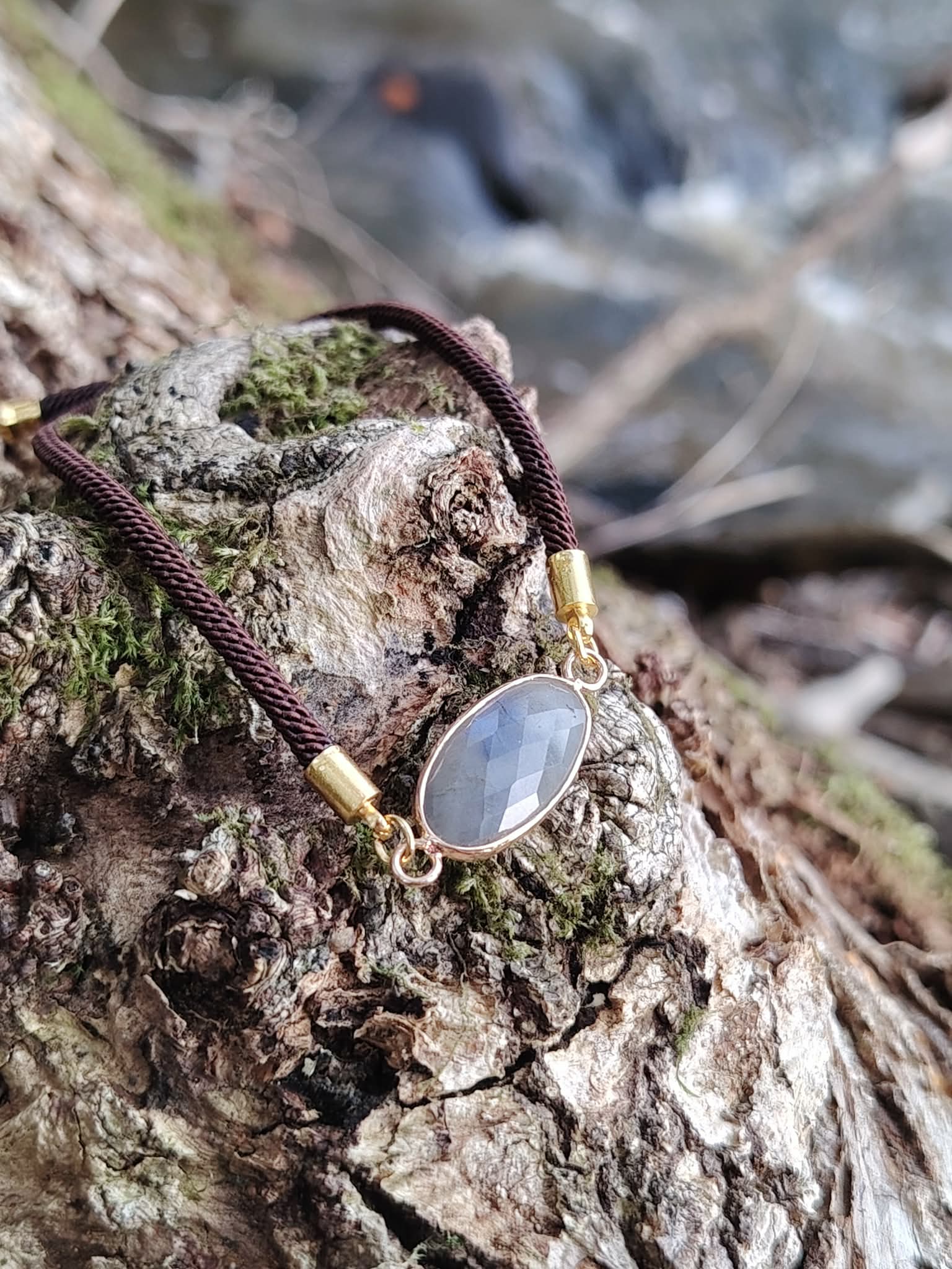 Bracelet médaillon ovale en labradorite sur bois moussu au bord d’un ruisseau, cordon brun et pierre facettée gris bleuté, calme mental et intuition.