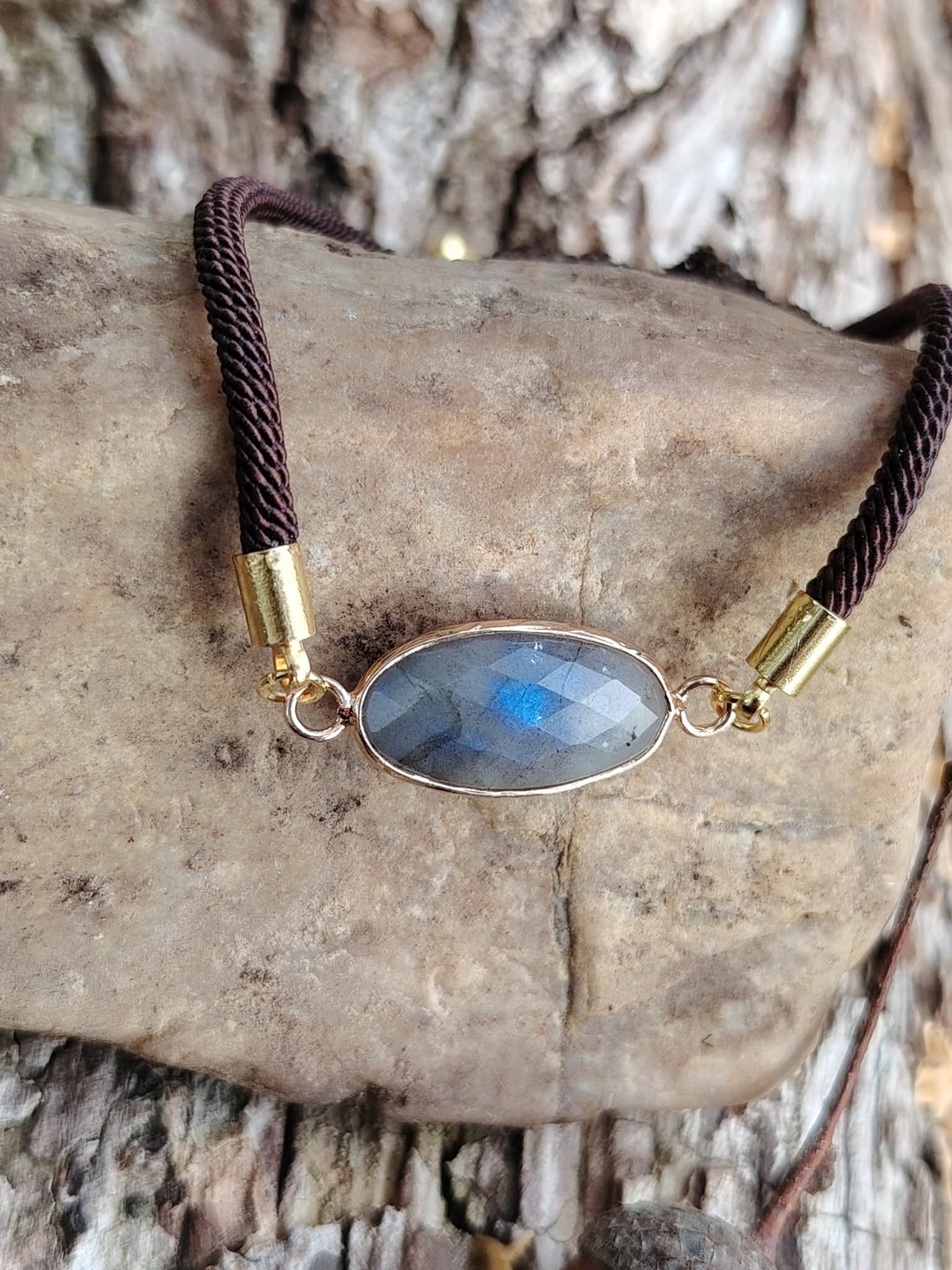 Bracelet médaillon ovale facetté en labradorite sur pierre claire contre une écorce en forêt, cordon brun et monture dorée, intuition et protection subtile.