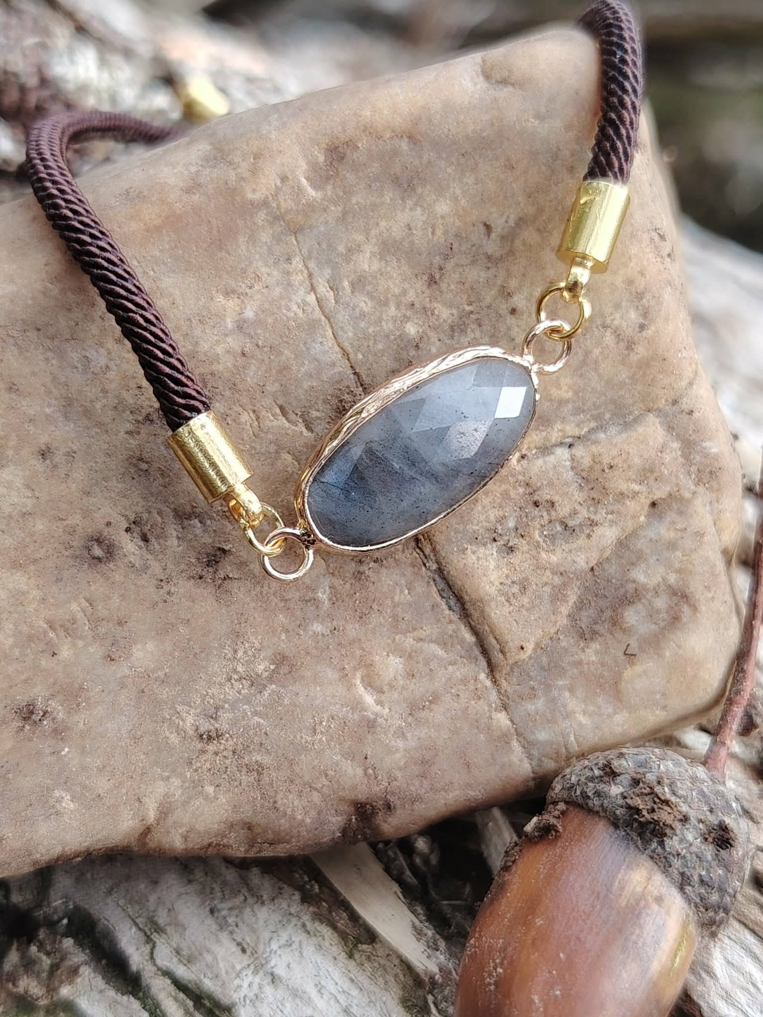 Zoom sur bracelet médaillon ovale en labradorite facettée posé sur pierre avec gland en décor naturel, cordon brun et monture dorée, intuition et stabilité émotionnelle.