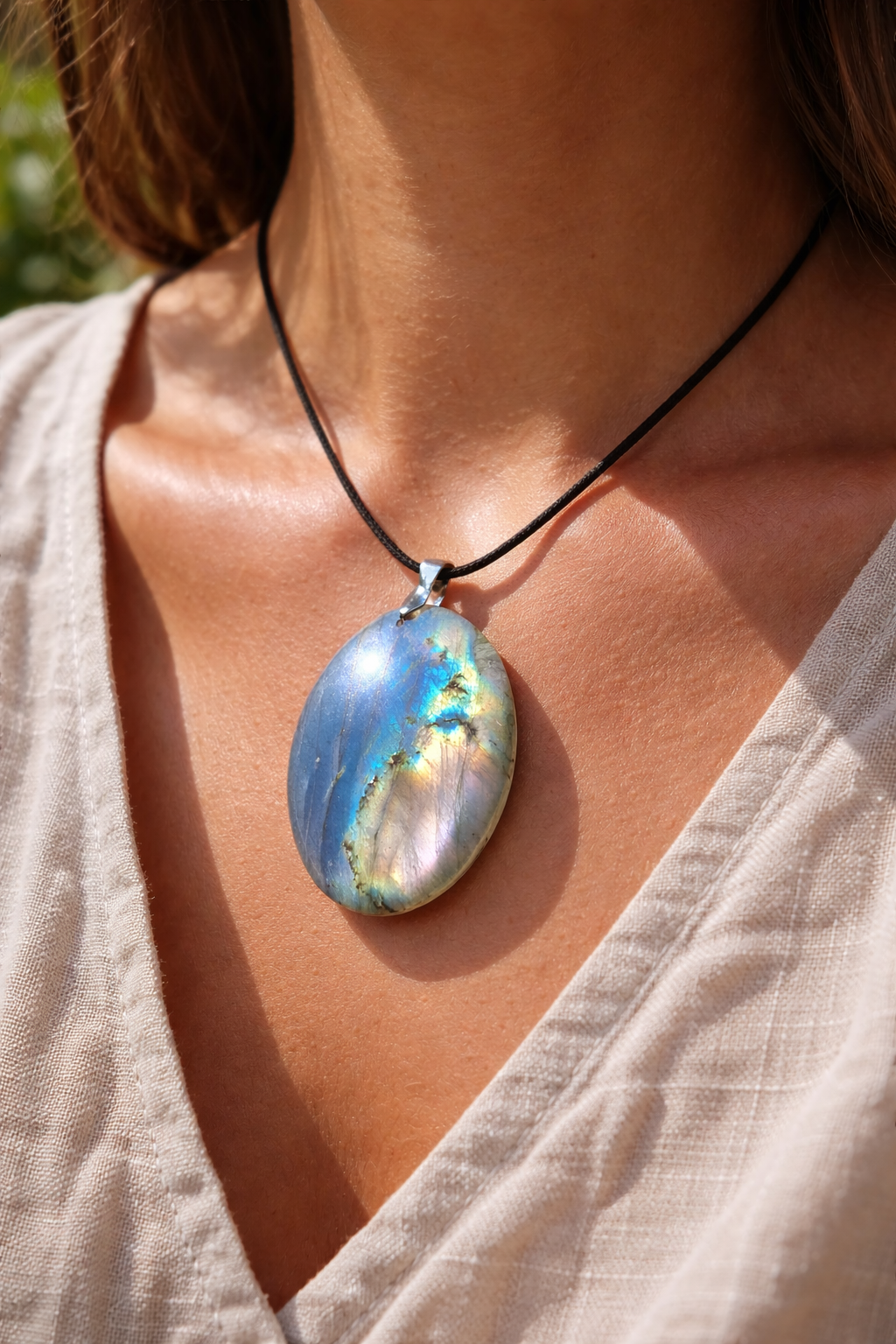 Pendentif ovale en labradorite bleue porté sur cordon noir, reflets bleu vert irisés sur peau en lumière naturelle, tenue beige et élégance protectrice.