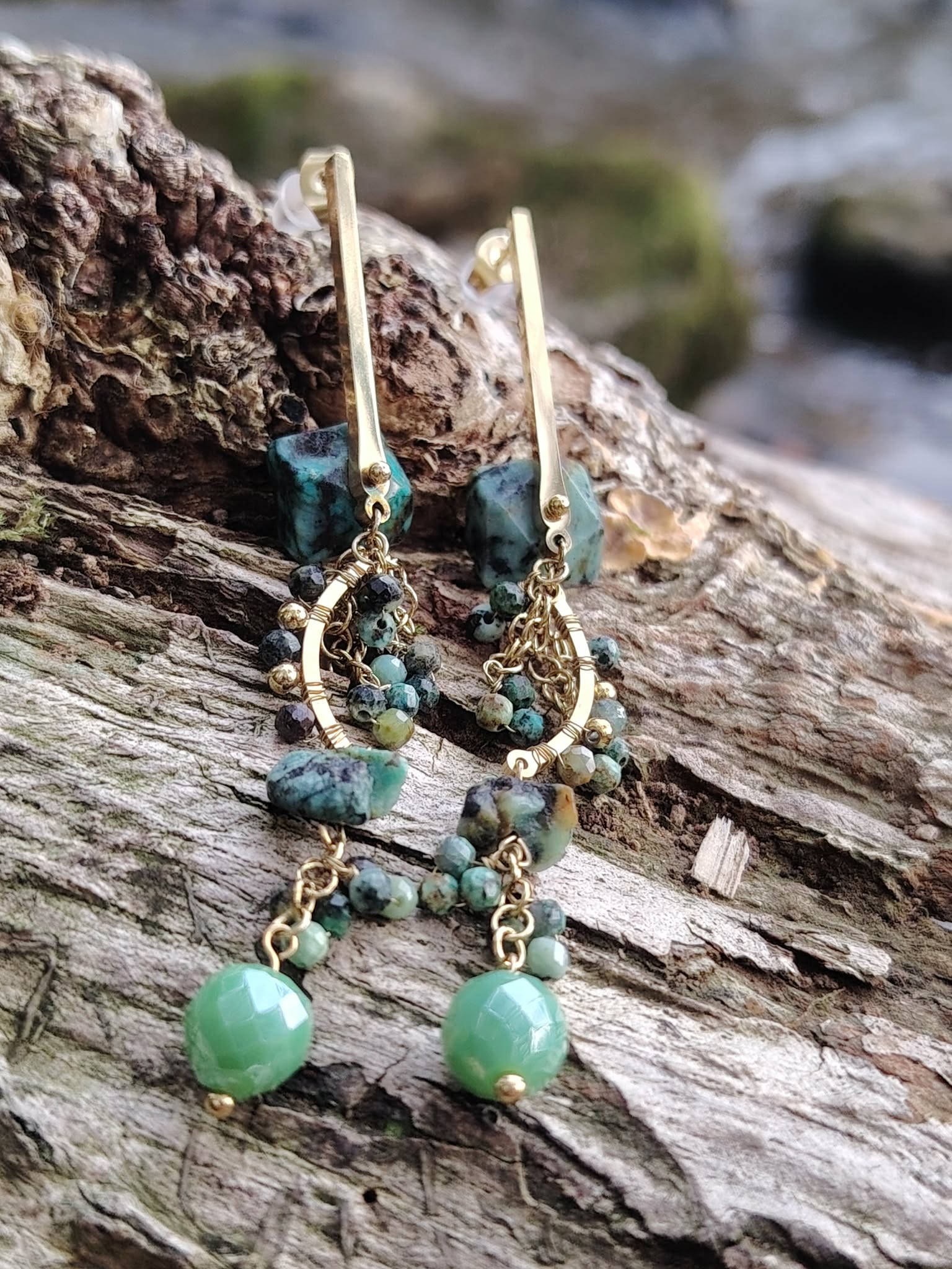 Boucles d’oreilles dorées pendantes en turquoise africaine sur bois texturé en forêt, perles bleu-vert marbrées et pampilles délicates, apaisement intérieur et expression fluide.
