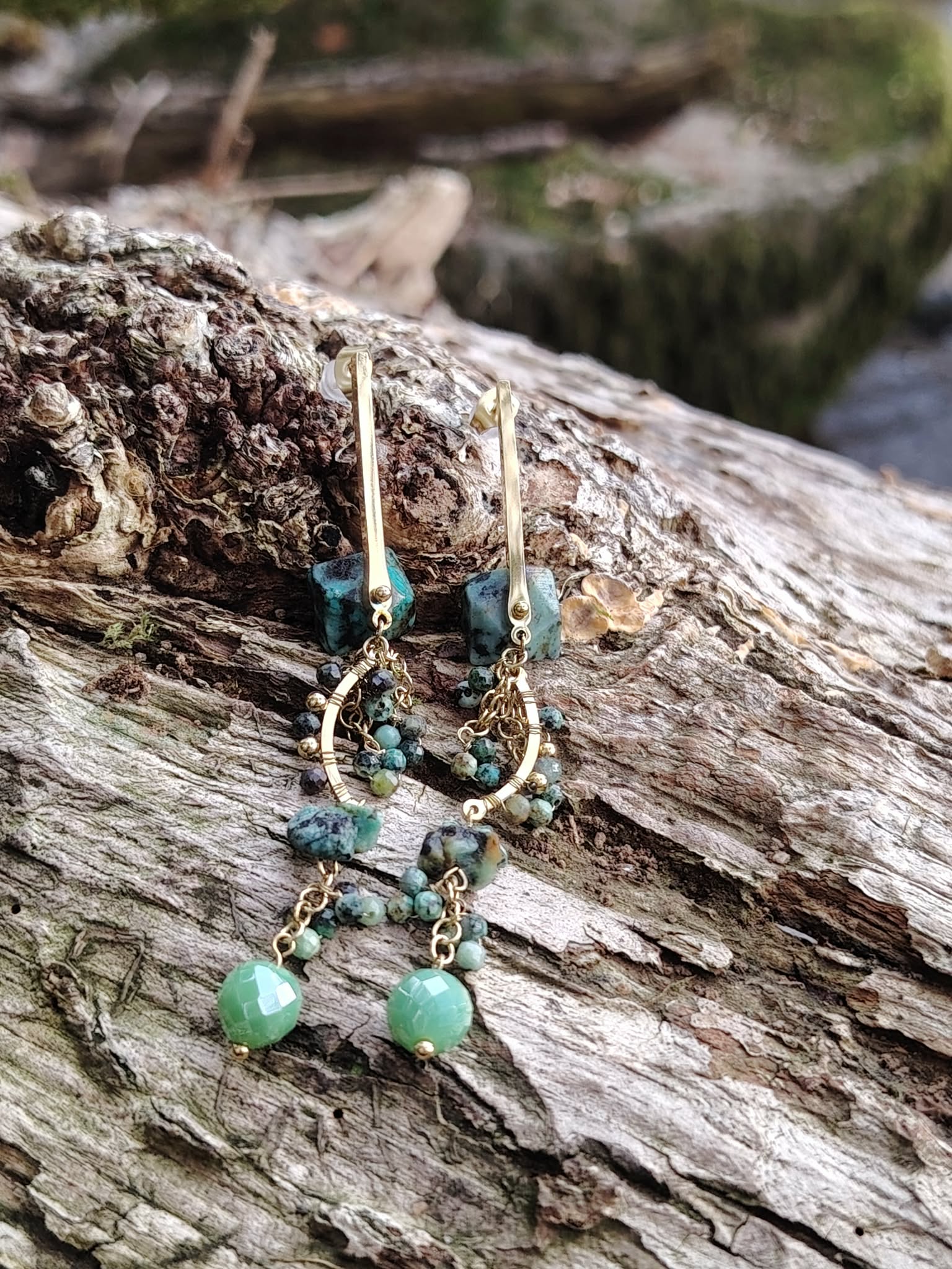 Boucles d’oreilles dorées turquoise africaine sur tronc en forêt, pierres naturelles bleu-vert et pampilles fines, douceur émotionnelle et allure bohème