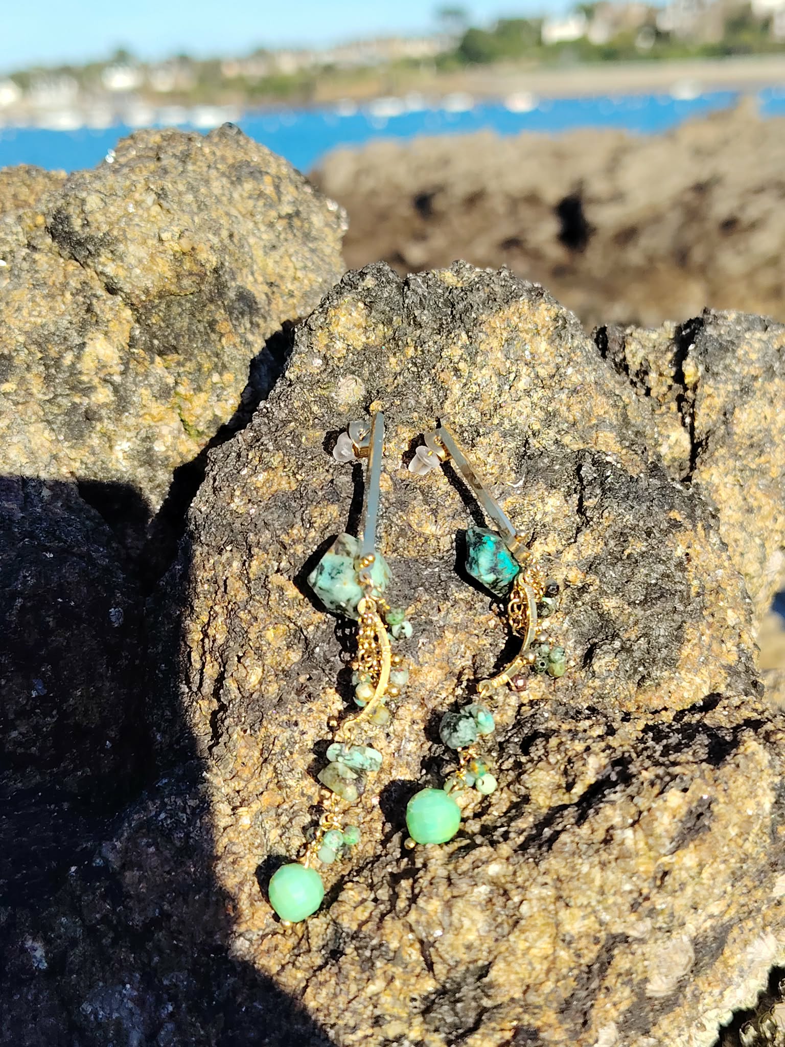 Boucles d’oreilles pendantes en turquoise africaine sur rocher au bord de mer en Bretagne, nuances bleu-vert marbrées et métal doré lumineux, communication apaisée et harmonie.