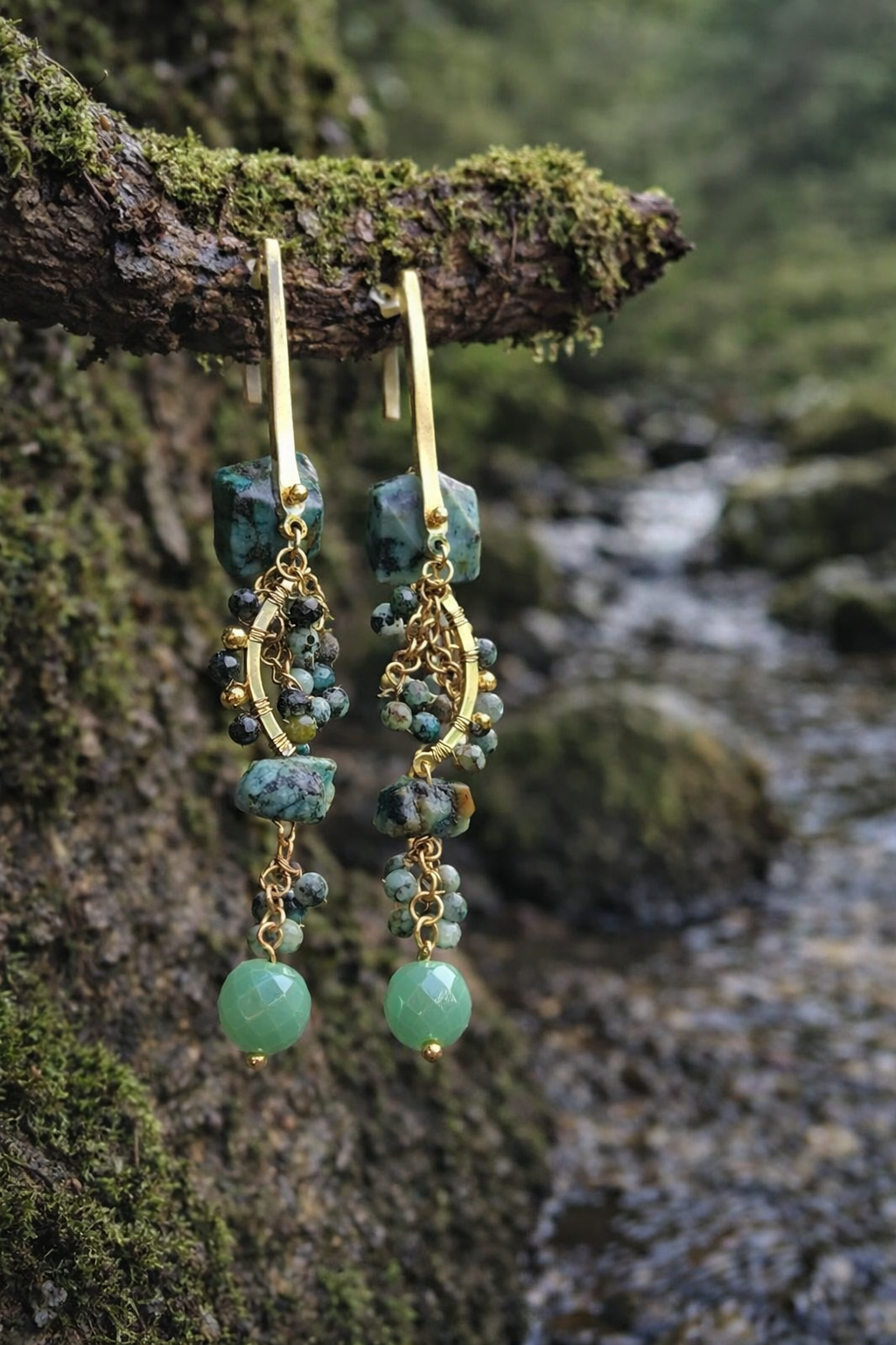 Boucles d’oreilles pendantes dorées avec pierres vertes style turquoise africaine suspendues sur branche moussue au bord d’un ruisseau, esprit bohème chic et élégance naturelle.