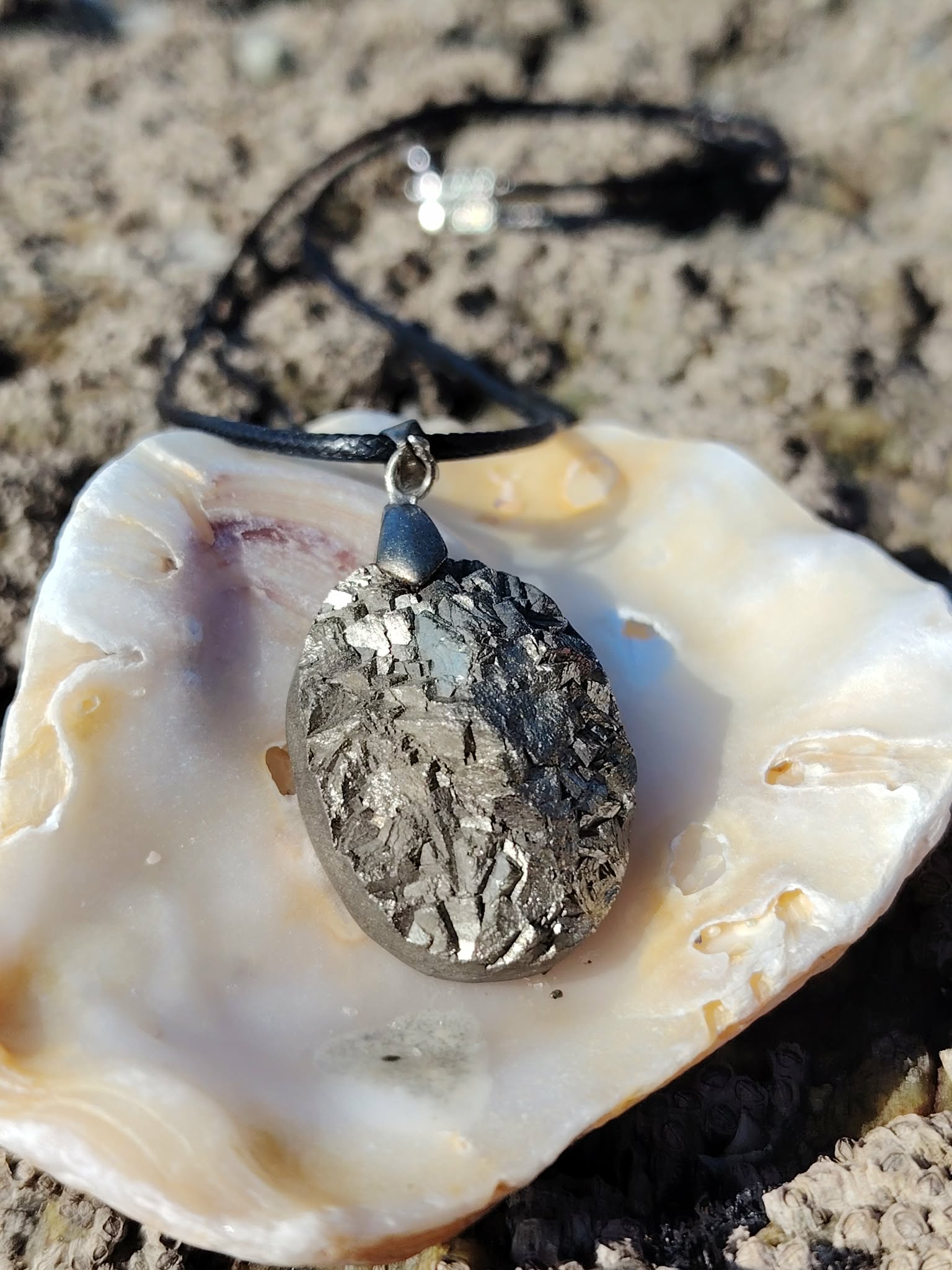 Pendentif pyrite naturelle sur coquillage blanc en extérieur en Bretagne, pierre ovale aux reflets métalliques avec cordon noir, symbole d’élan, de courage et de protection.