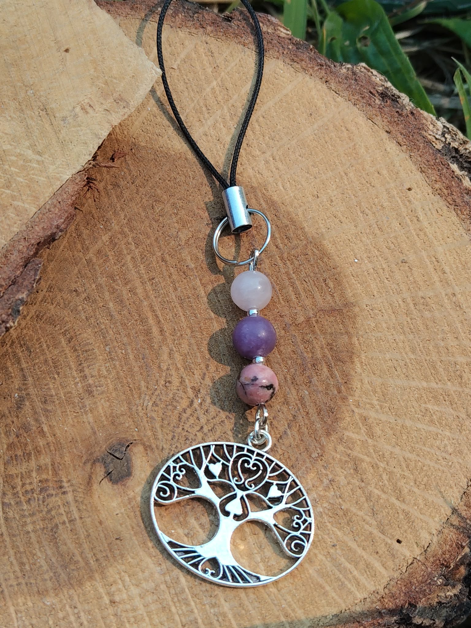 Bijou de téléphone arbre de vie en kunzite, lépidolite et rhodonite sur bois naturel avec feuillage en fond, création Magic Gaïa aux tons rose et mauve.