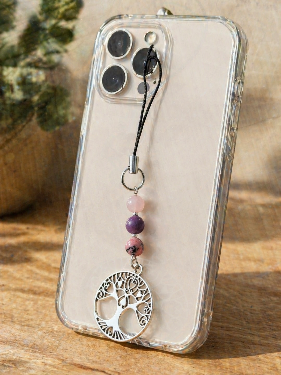 Bijou de téléphone arbre de vie avec kunzite, lépidolite et rhodonite accroché à une coque transparente sur support en bois, lumière naturelle et fond végétal flou.