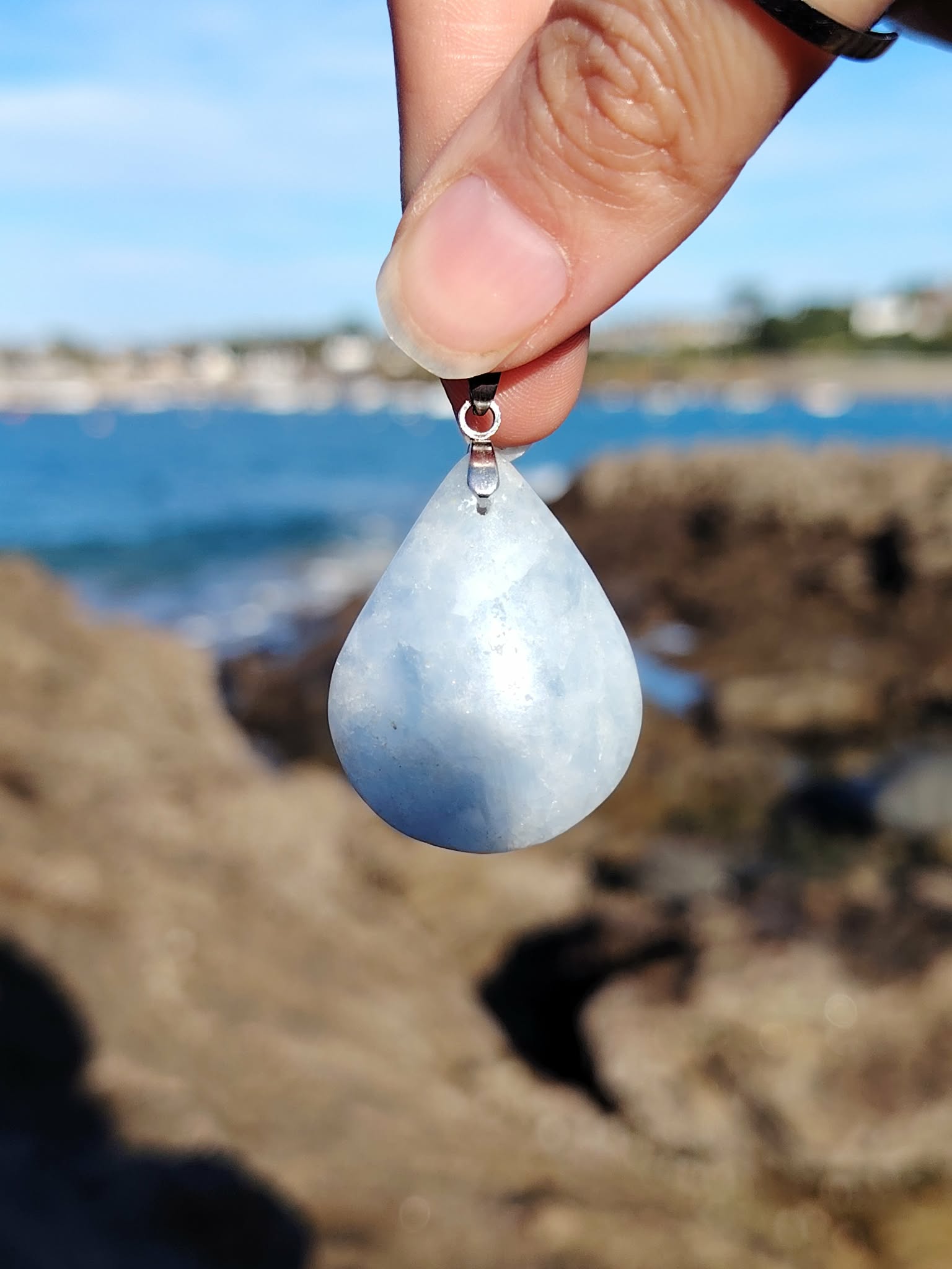 Pendentif calcite bleue naturelle posé sur bois en Bretagne avec arrière-plan de nature, pierre goutte bleu pastel, sérénité et lâcher-prise.
