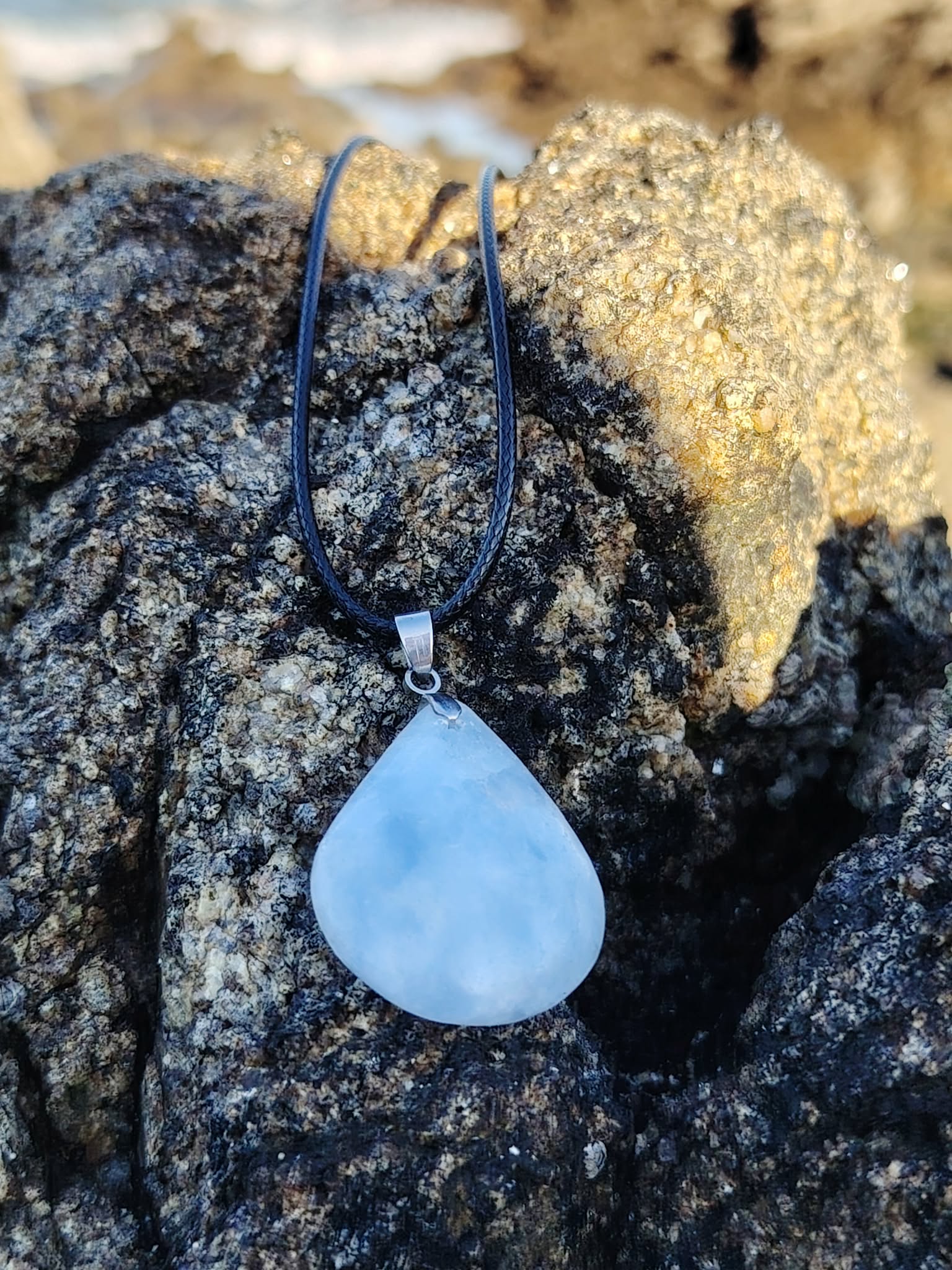 Pendentif calcite bleue naturelle en goutte sur bois en Bretagne avec décor de bord de mer, pierre bleu laiteux polie sur cordon noir, apaisement et communication douce.